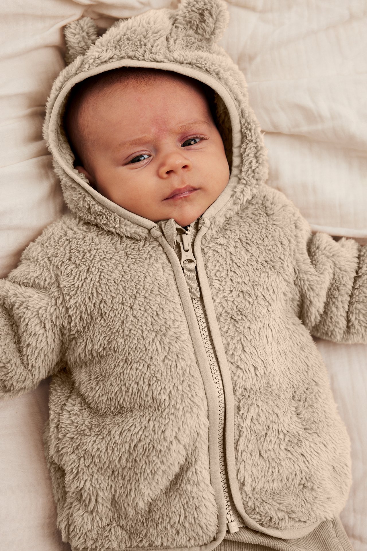 Ylhäältä näkymä vauvasta beigessä pörröisessä hupputakissa korvilla ja vetoketjulla, beiget housut.