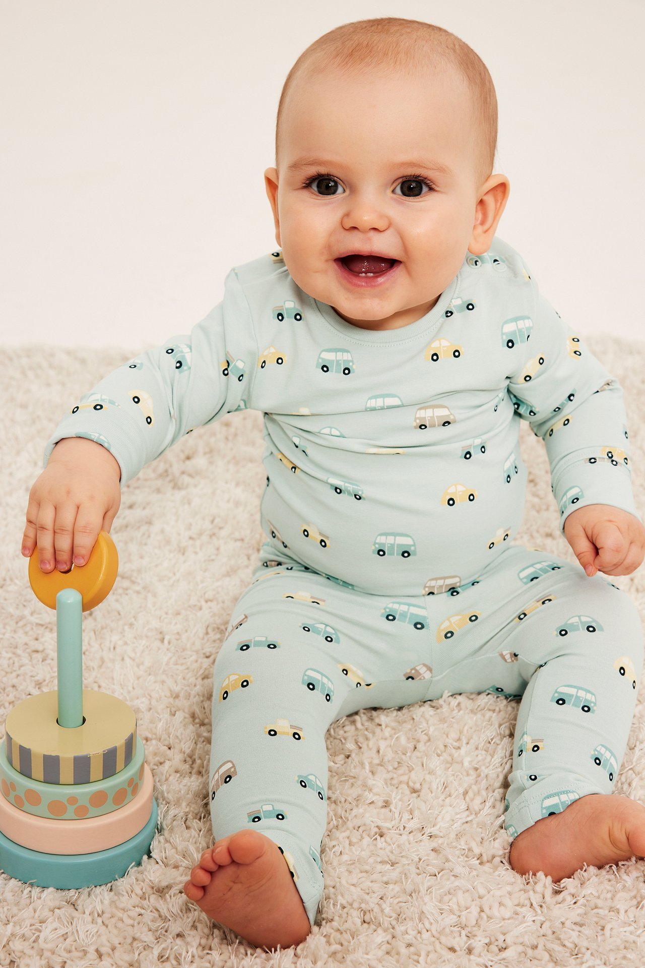 Smilende baby i lyseblå todelt pysjamas med biltrykk, holder treleke.