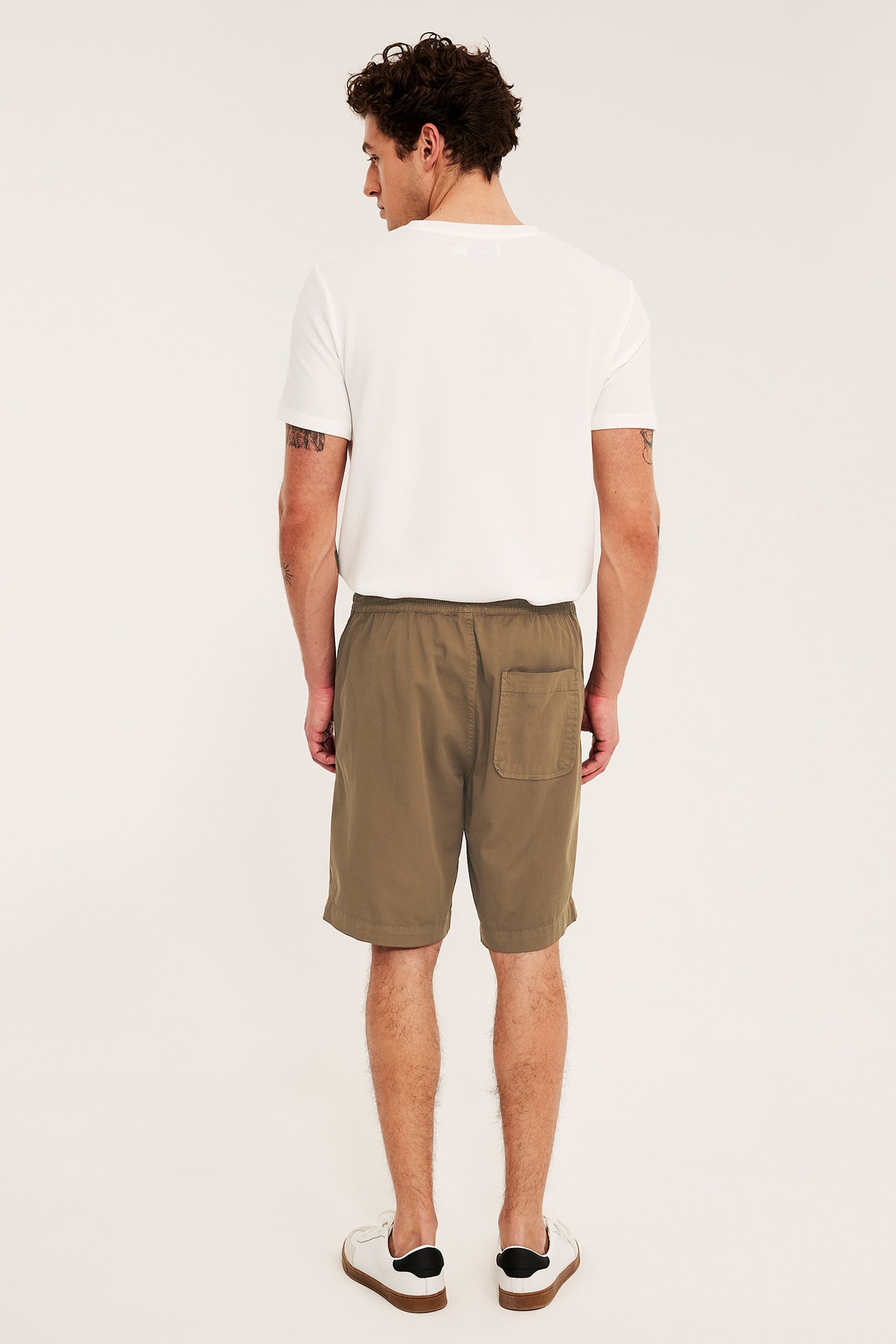 Takaapäin: Mies ruskeissa väljissä puuvillatwillishortseissa, joustovyötäröllä, valkoinen t-paita ja valkoiset tennarit.