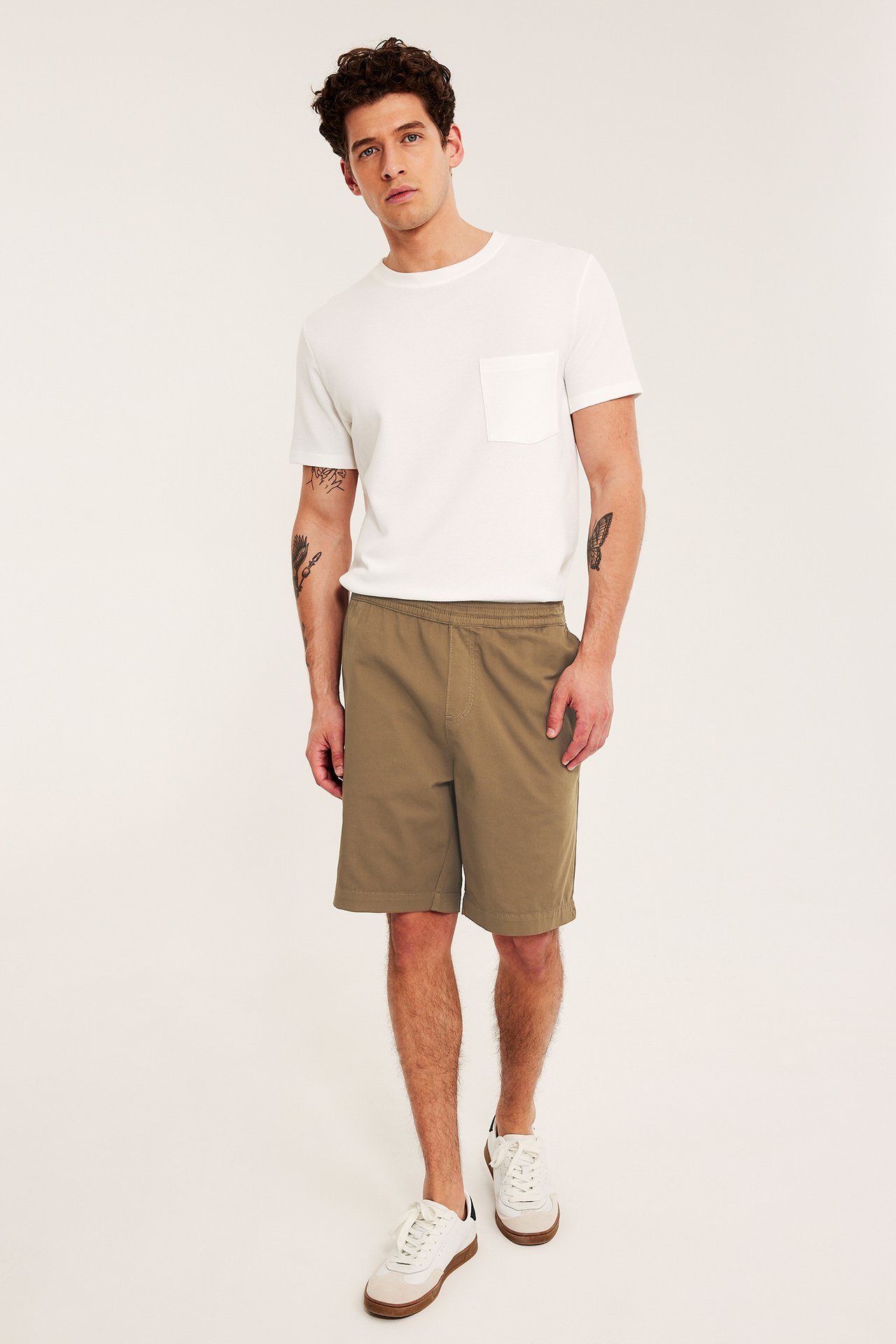 Framifrån, en man i bruna lösa bomullstwillshorts, en vit t-shirt och vita sneakers.