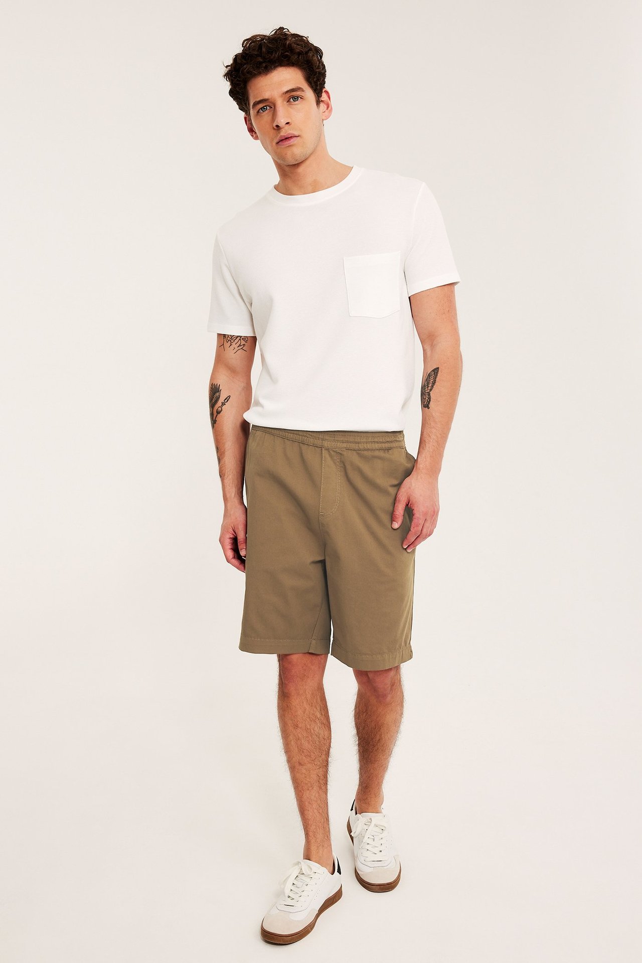 Framifrån, en man i bruna lösa bomullstwillshorts, en vit t-shirt och vita sneakers.