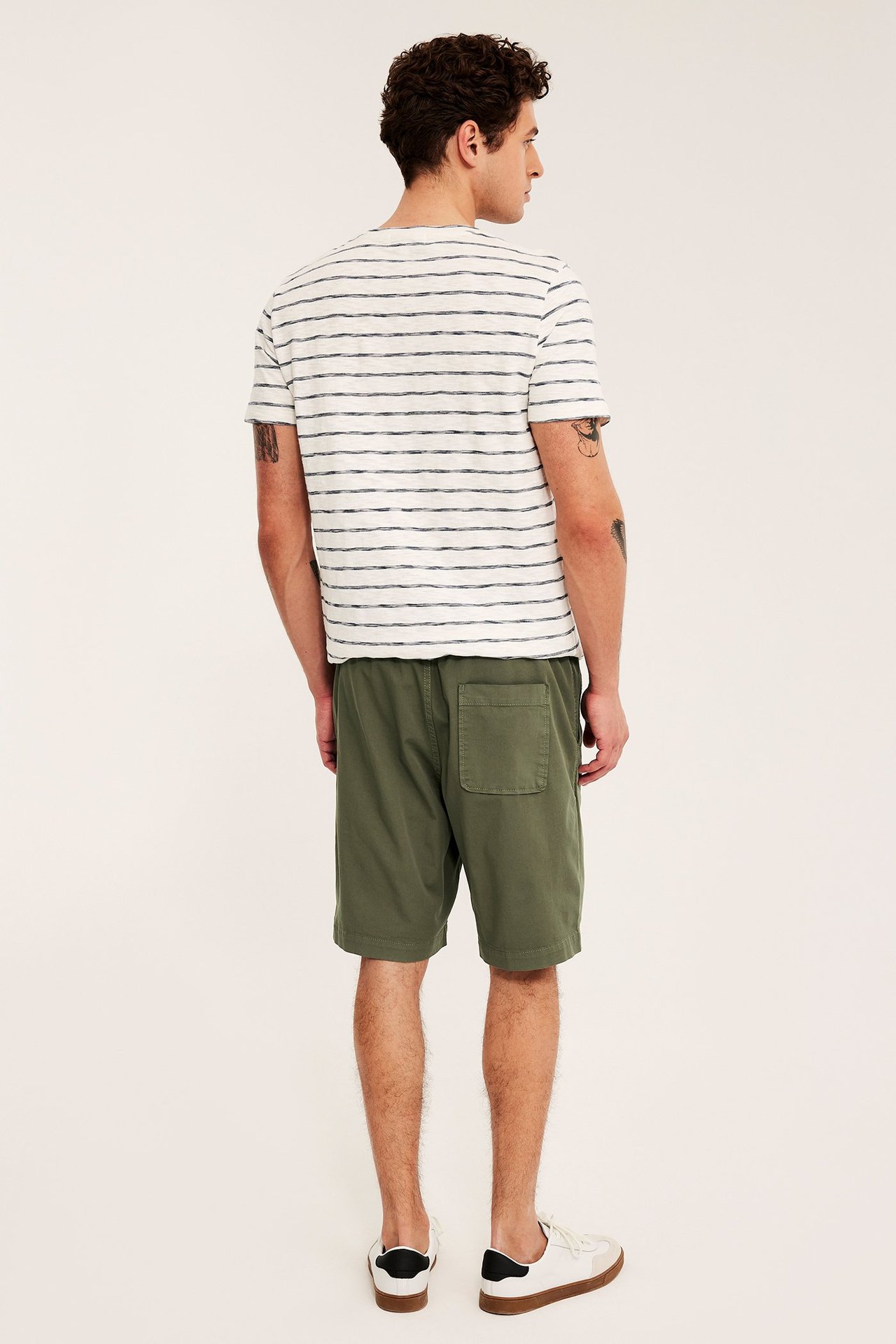 Mies takaa, yllään vihreät väljät puuvillatvillishortsit, raidallinen t-paita ja valkoiset tennarit.