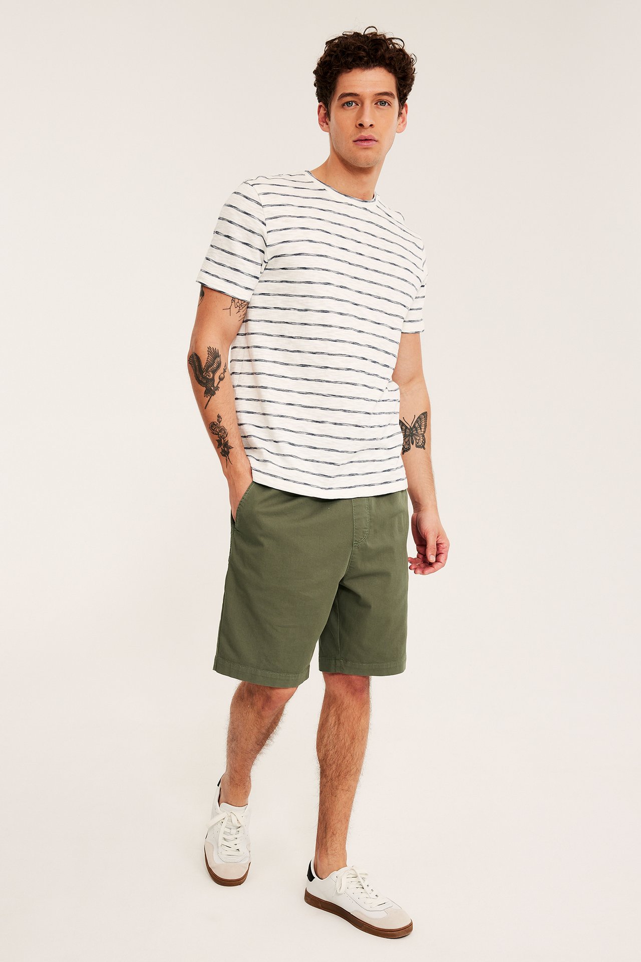 Forfra: mann i grønne loose fit bomullstwillshorts, en hvitstripet t-skjorte og hvite joggesko.