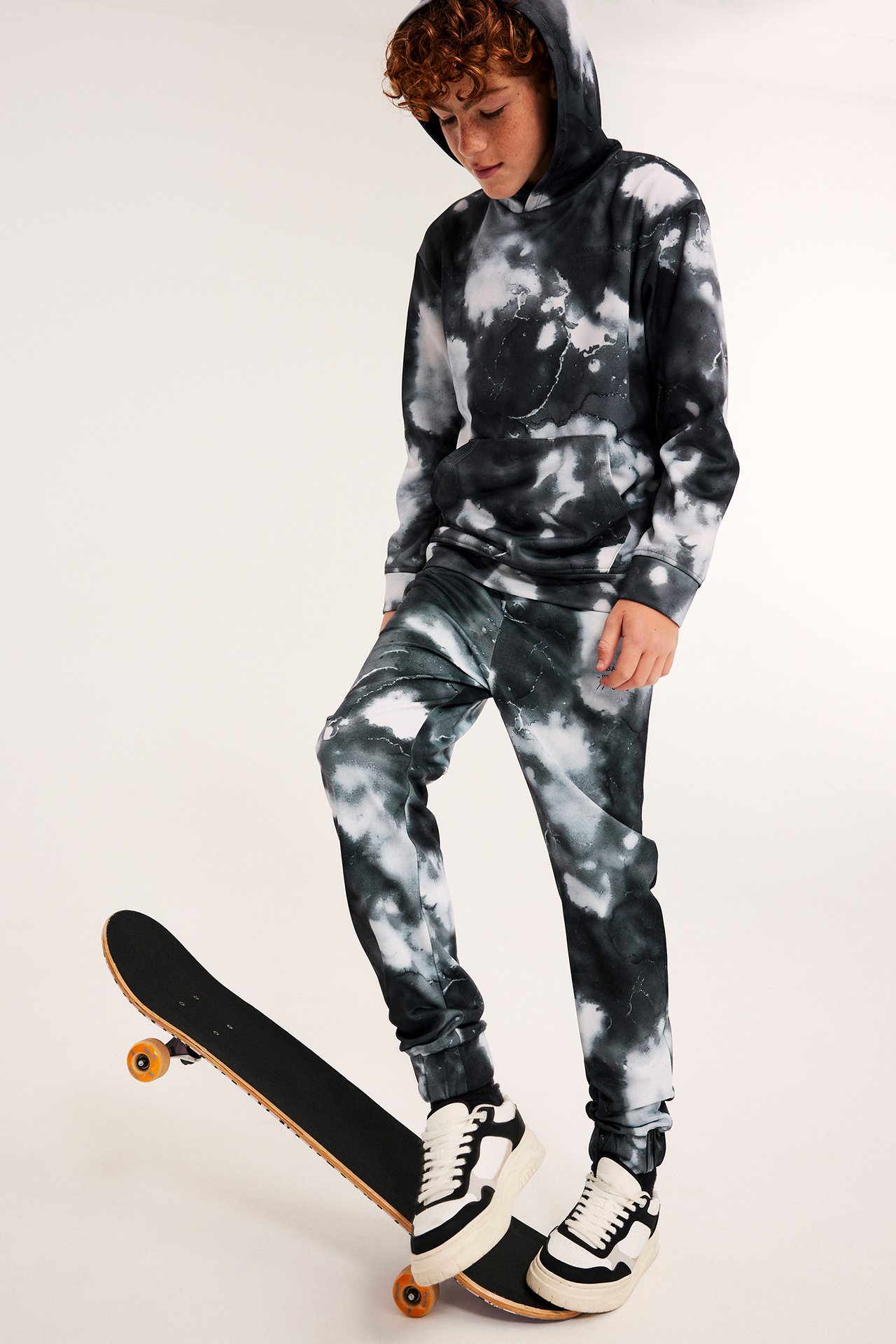 Forfra, et barn i et svart og hvitt tie-dye hettegenser og joggebuksesett, fot på skateboard.