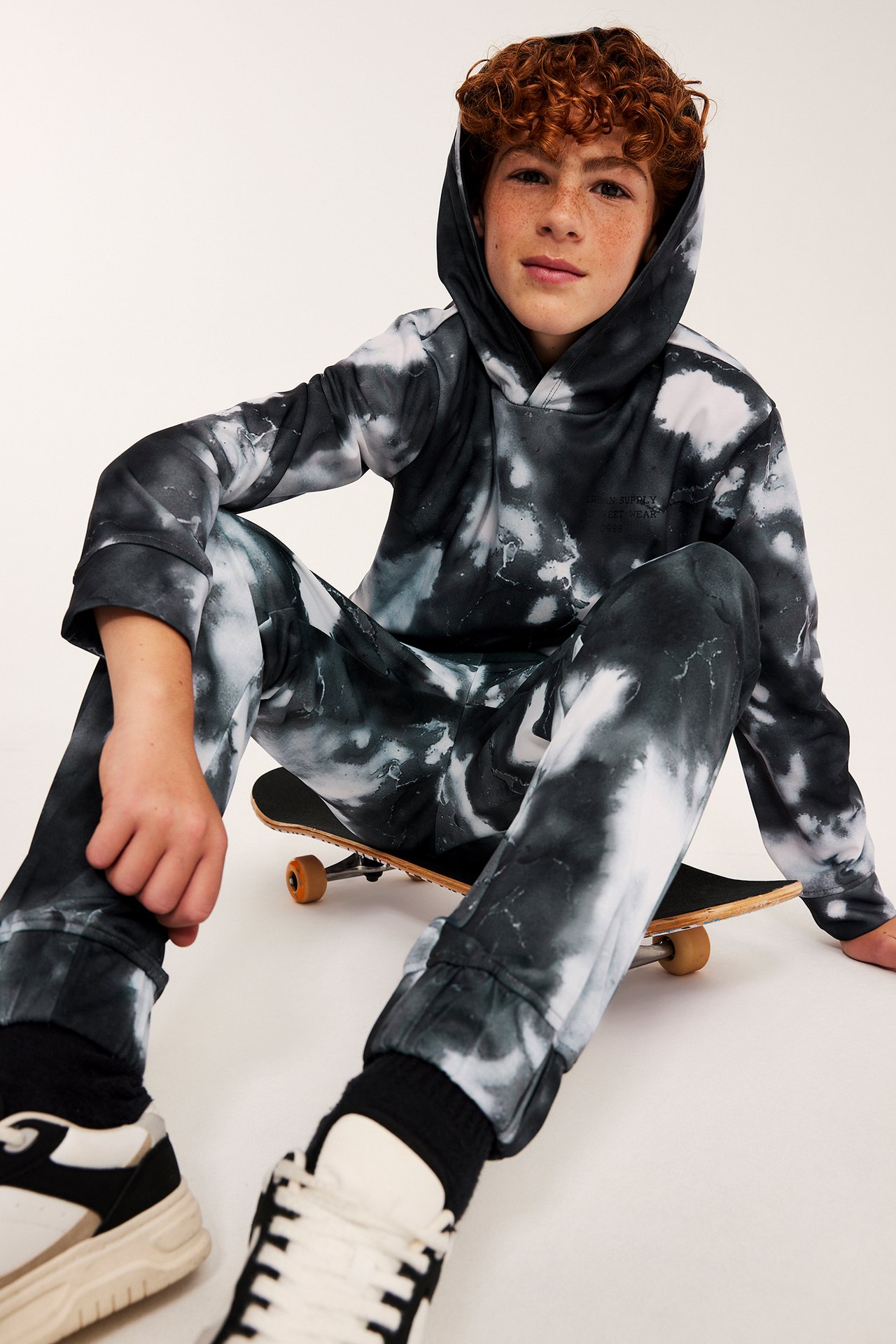 Widok z przodu dziecka w czarno-białym dresie tie-dye, siedzi ze deskorolką i sneakersami.