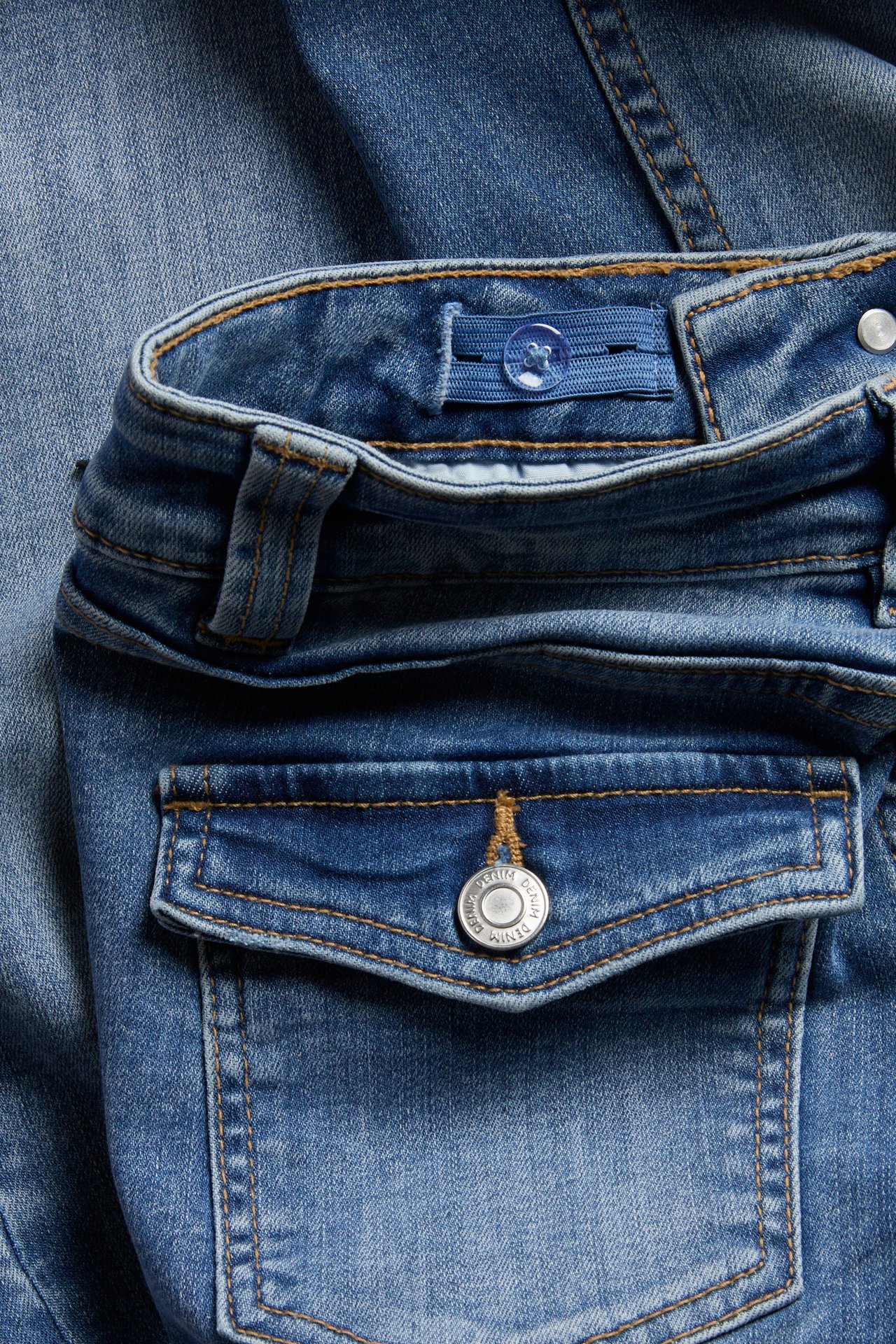 Närbild på barns ljusblå denimjeans, visar justerbar linning och en ficka med silverknapp.