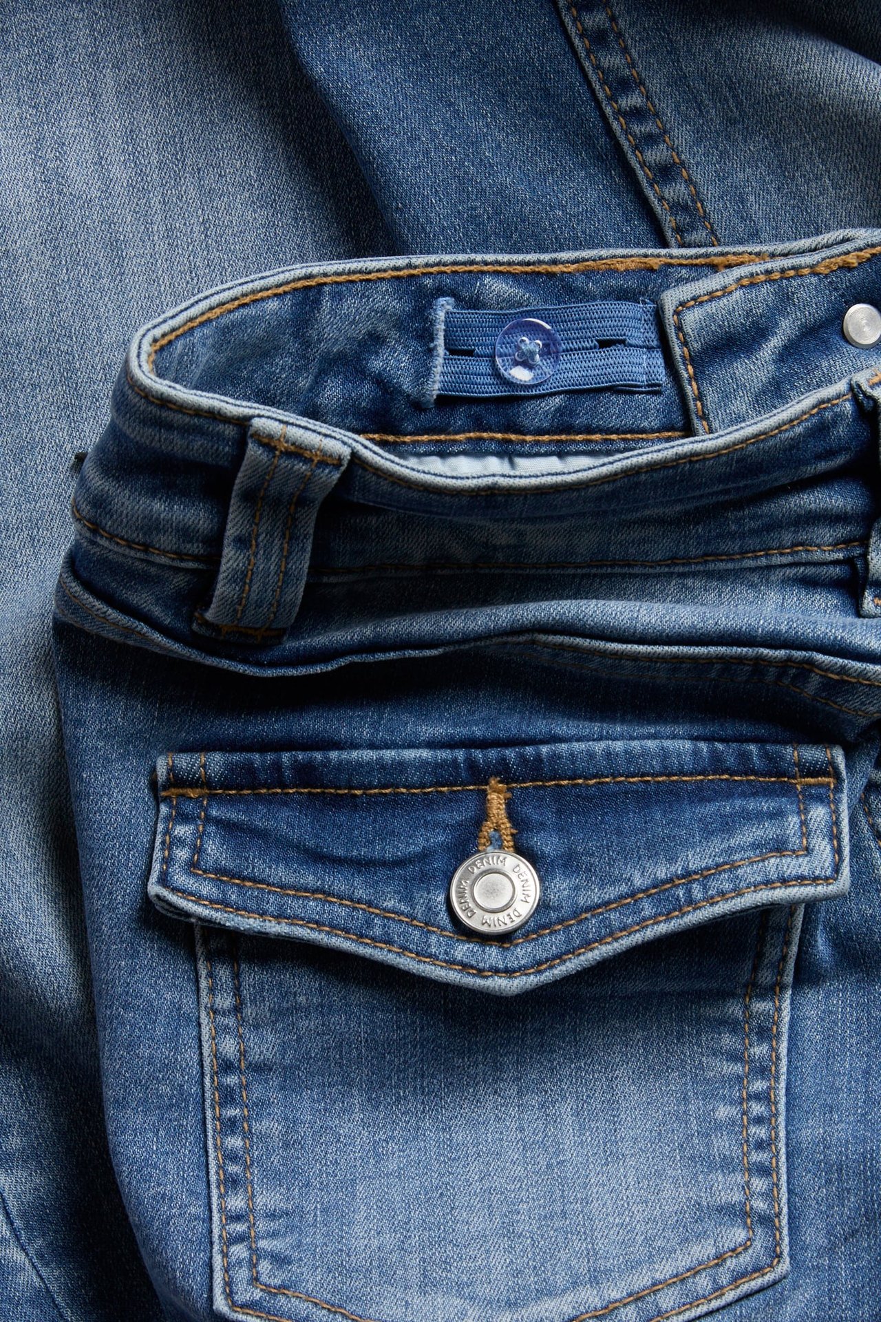 Zbliżenie na jasnoniebieskie jeansy dziecięce z denimu, widoczny regulowany pas i kieszeń z klapką z guzikiem.