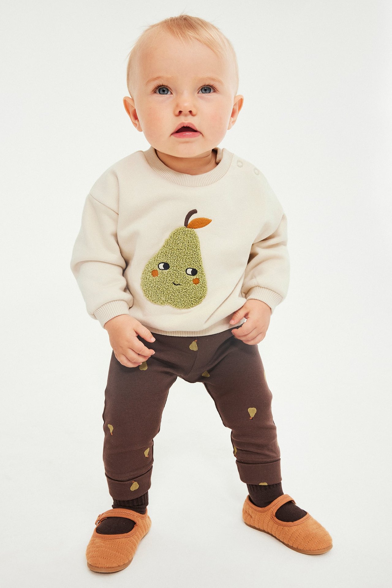 Frontbilde av baby i kremfarget genser med teksturert grønn pære, brune pæremønstrede leggings og brune sko.