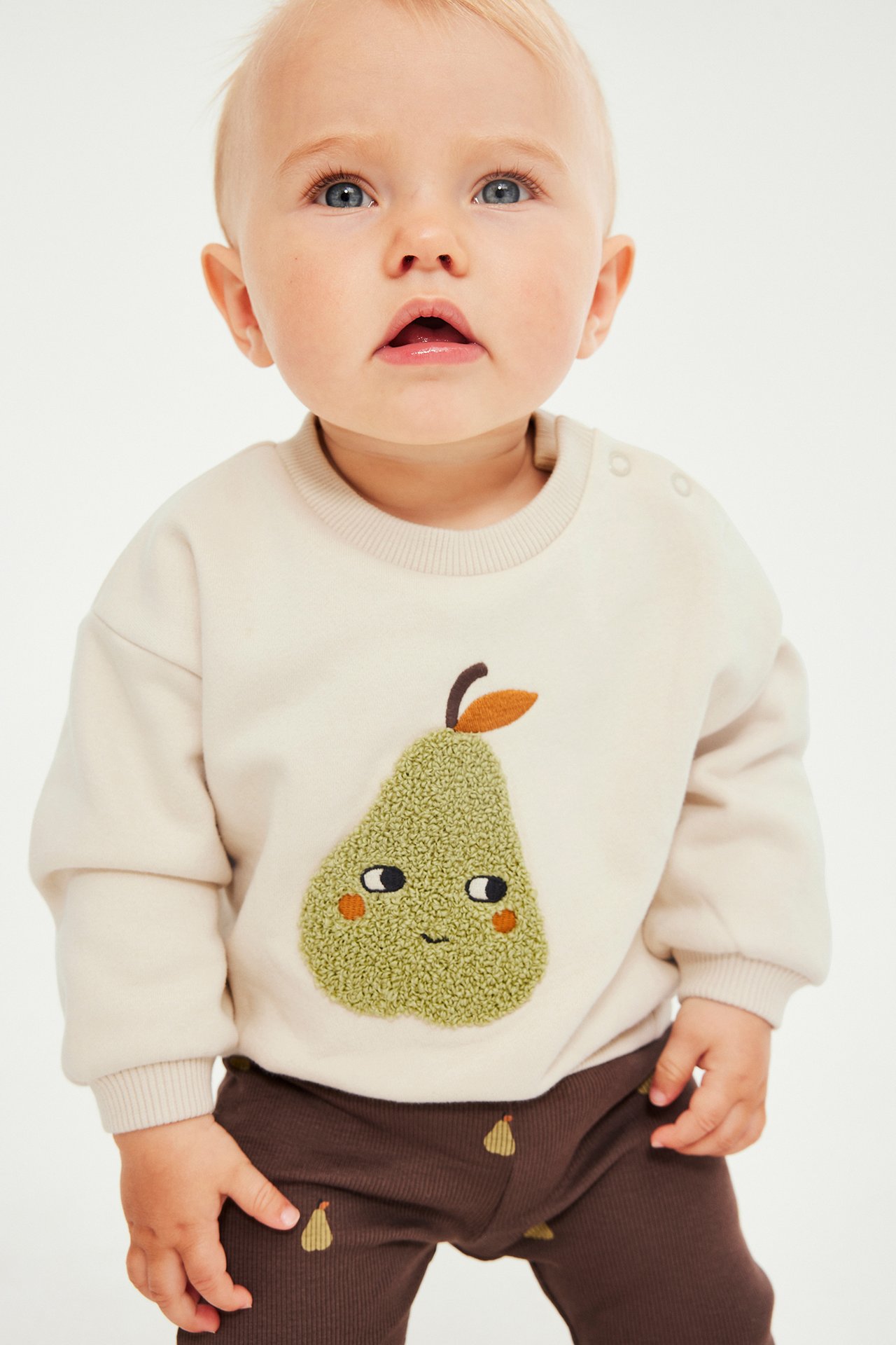 Frontbilde av baby i beige genser med pære-applikasjon og brune ribbestrikkede leggings.