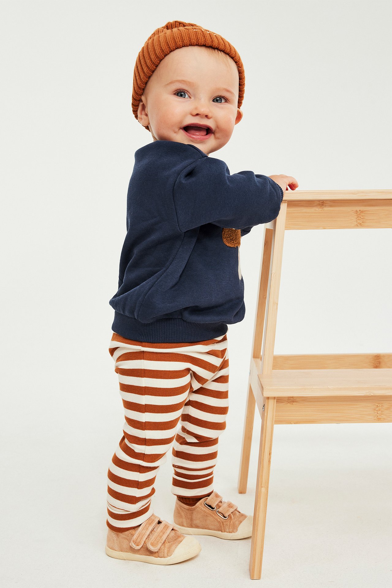 Baby i marinblå tröja, rostrandiga leggings, rostbrun mössa och beiga sneakers.