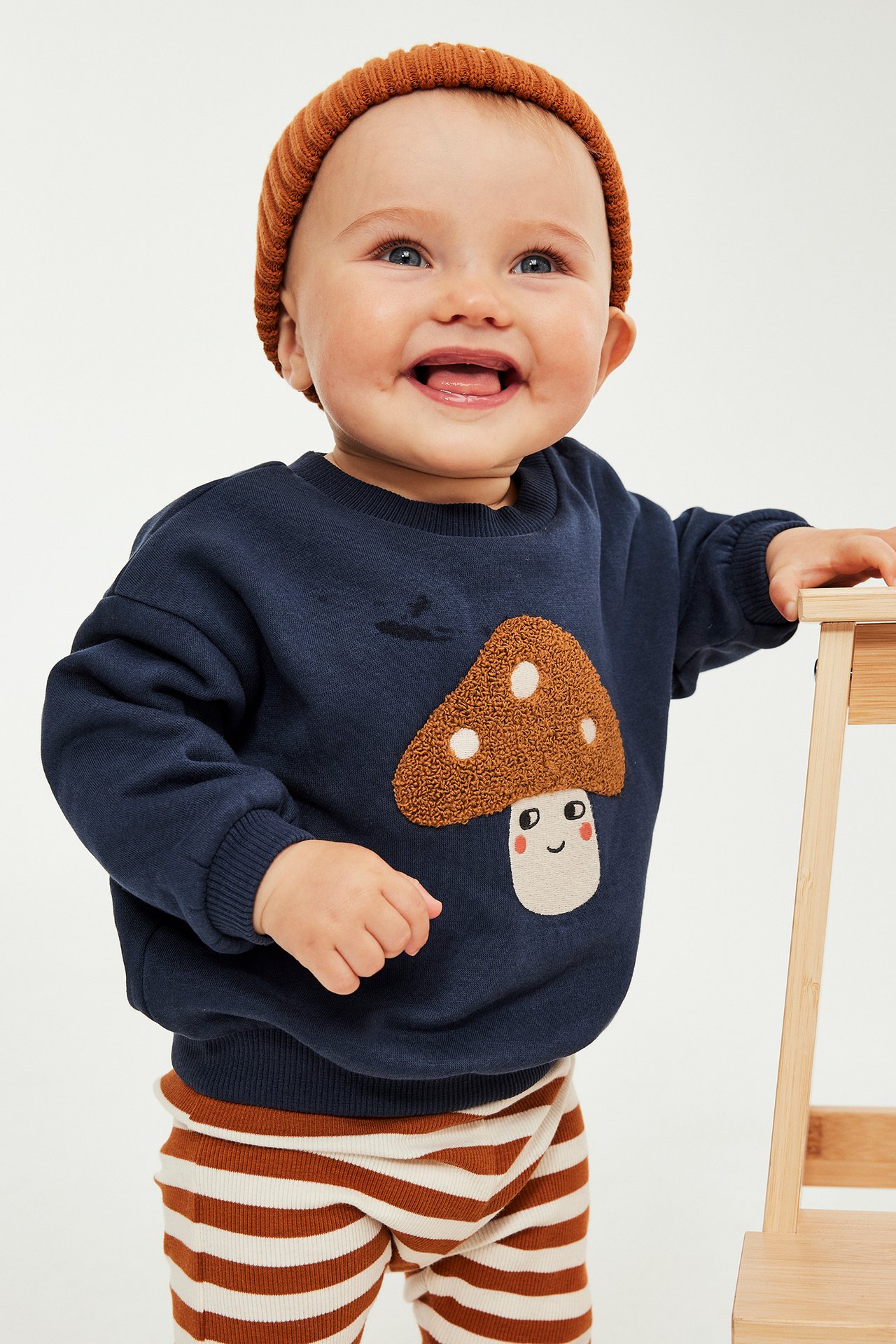 Baby i marinblå svamptröja, brun mössa och randiga ribbade leggings, håller pall.