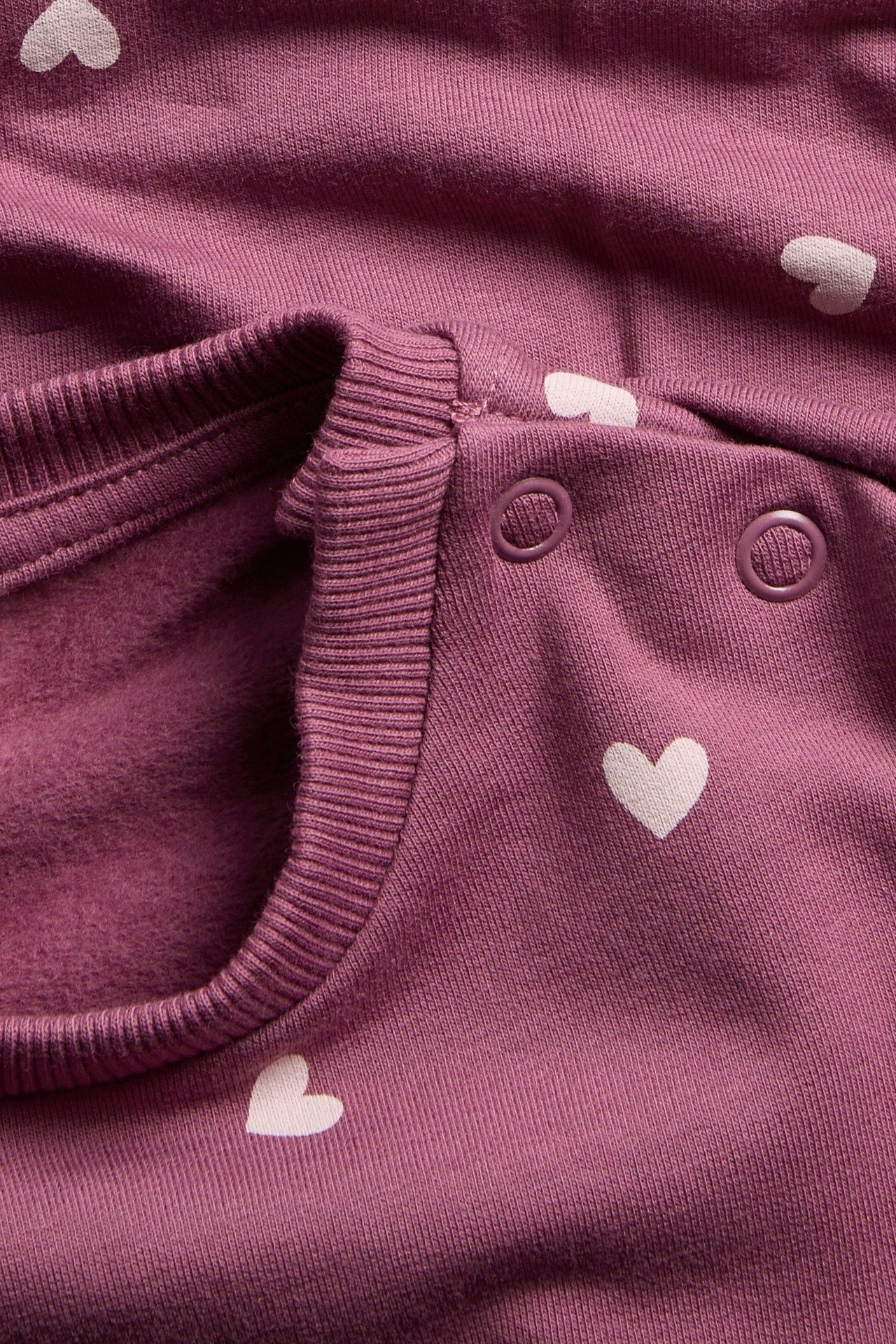 Närbild på en lila långärmad babytröja med vita hjärtan och tryckknappar på axeln.