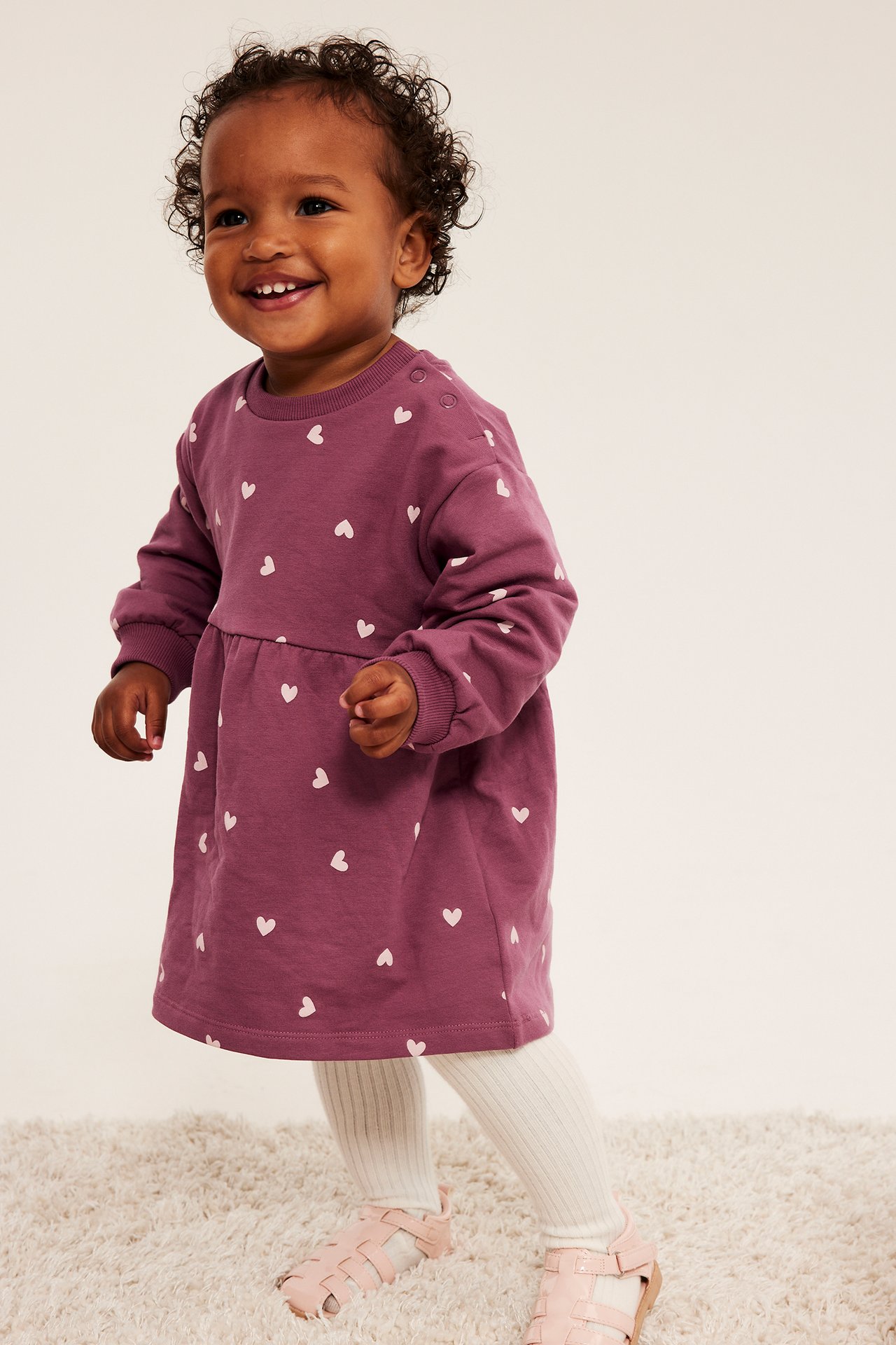 Framifrån: leende baby i dammrosa klänning med hjärtan, vita strumpbyxor och rosa sandaler.