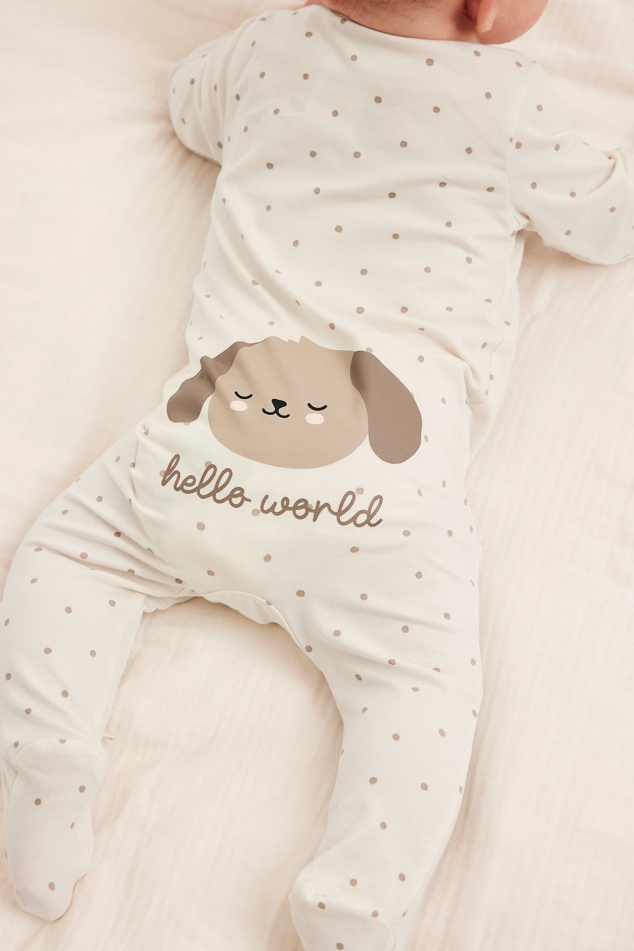 Tył niemowlęcia w białym śpiochu w kropki z brązową twarzą psa i napisem 'hello world'.