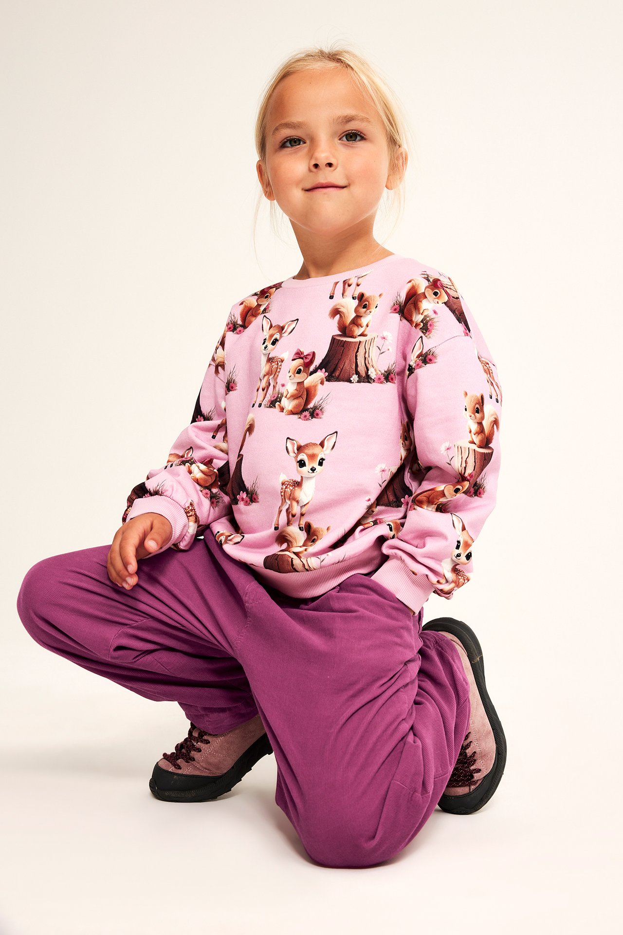 Framifrån, ett barn i rosa djurmönstrad sweatshirt, lila mjukisbyxor och bruna snörskor.