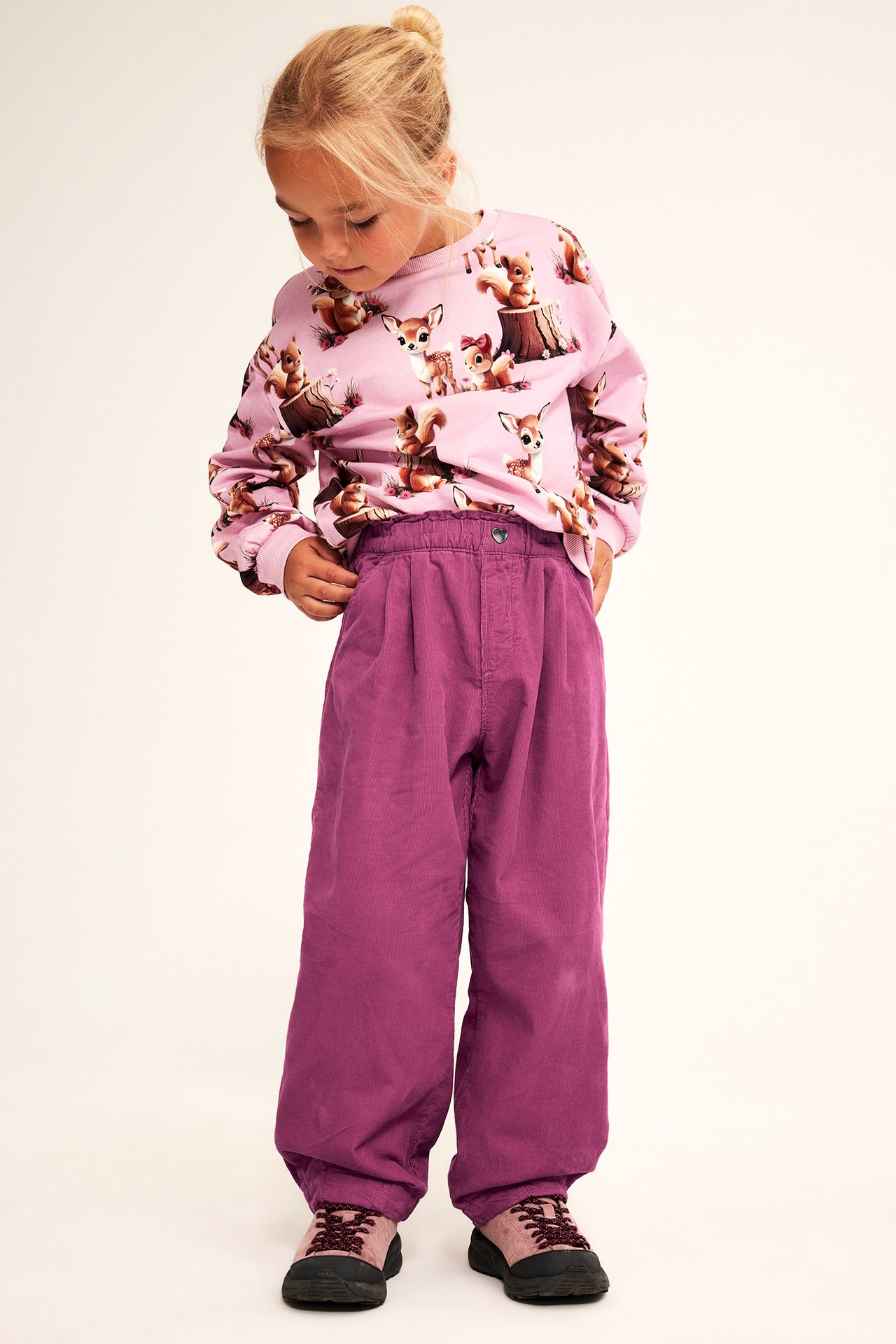 Frontvy av ett barn i lila vida plisserade byxor, en rosa långärmad tröja med djurmönster och sneakers.