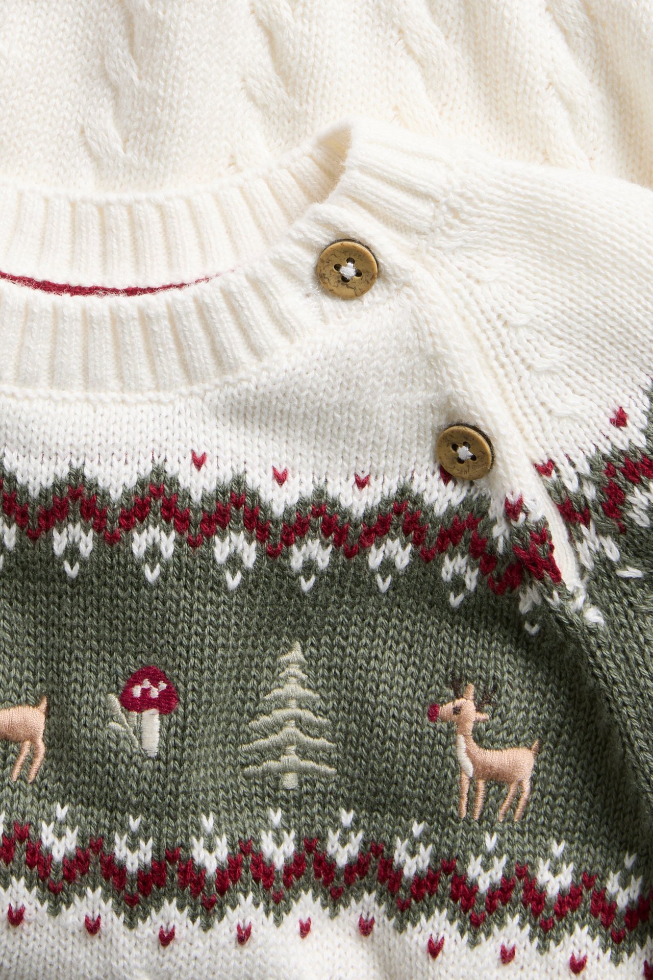 Närbild på en stickad babytröja i gräddvitt/olivgrönt med Fair Isle-mönster, skogsmotiv och träknappar.