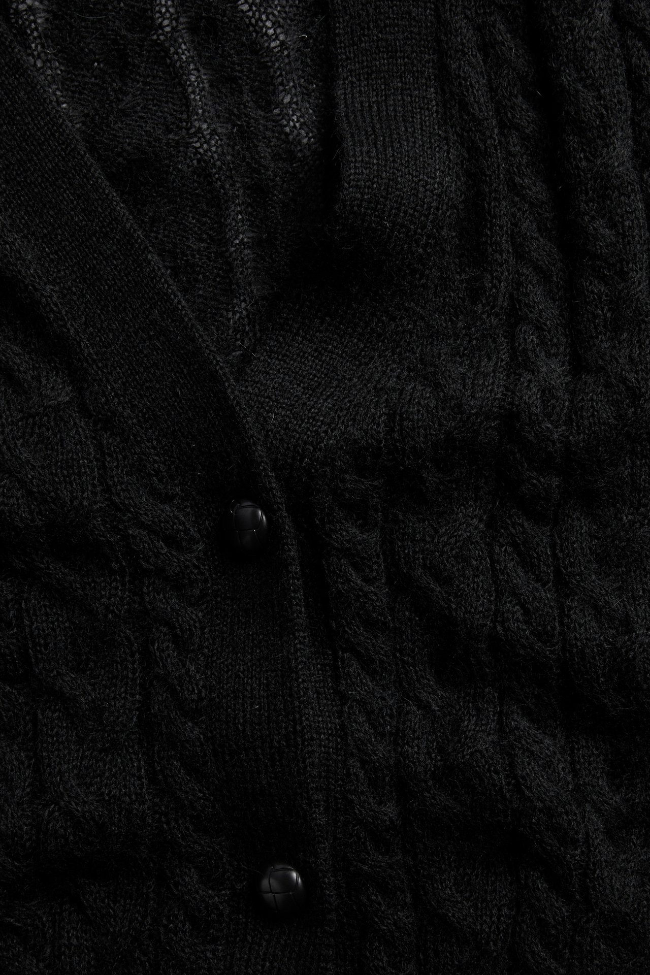 Nærbilde av en svart flettet cardigan med ribbestrikket stolpe og to runde svarte knapper.