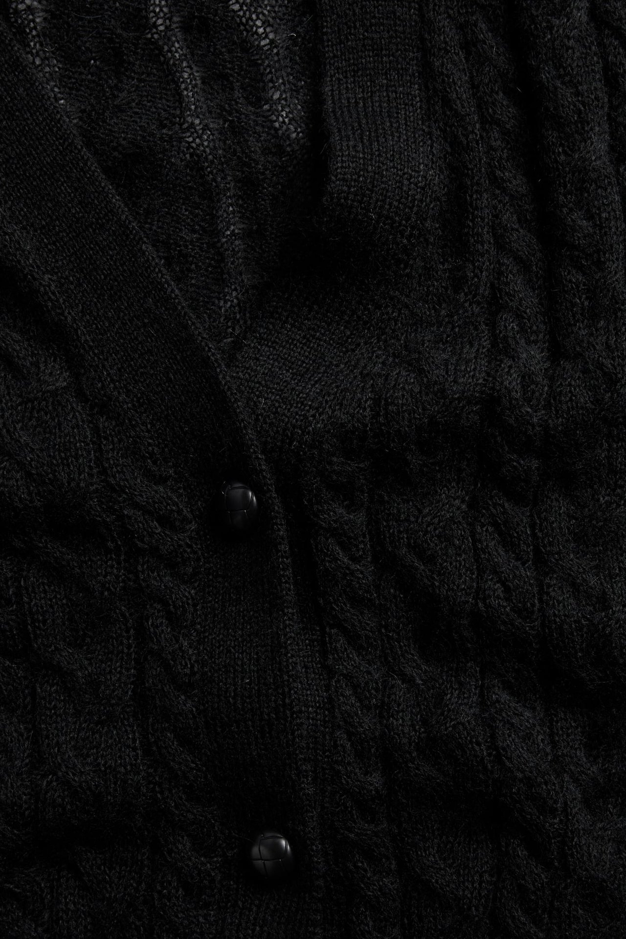 Nærbilde av en svart flettet cardigan med ribbestrikket stolpe og to runde svarte knapper.