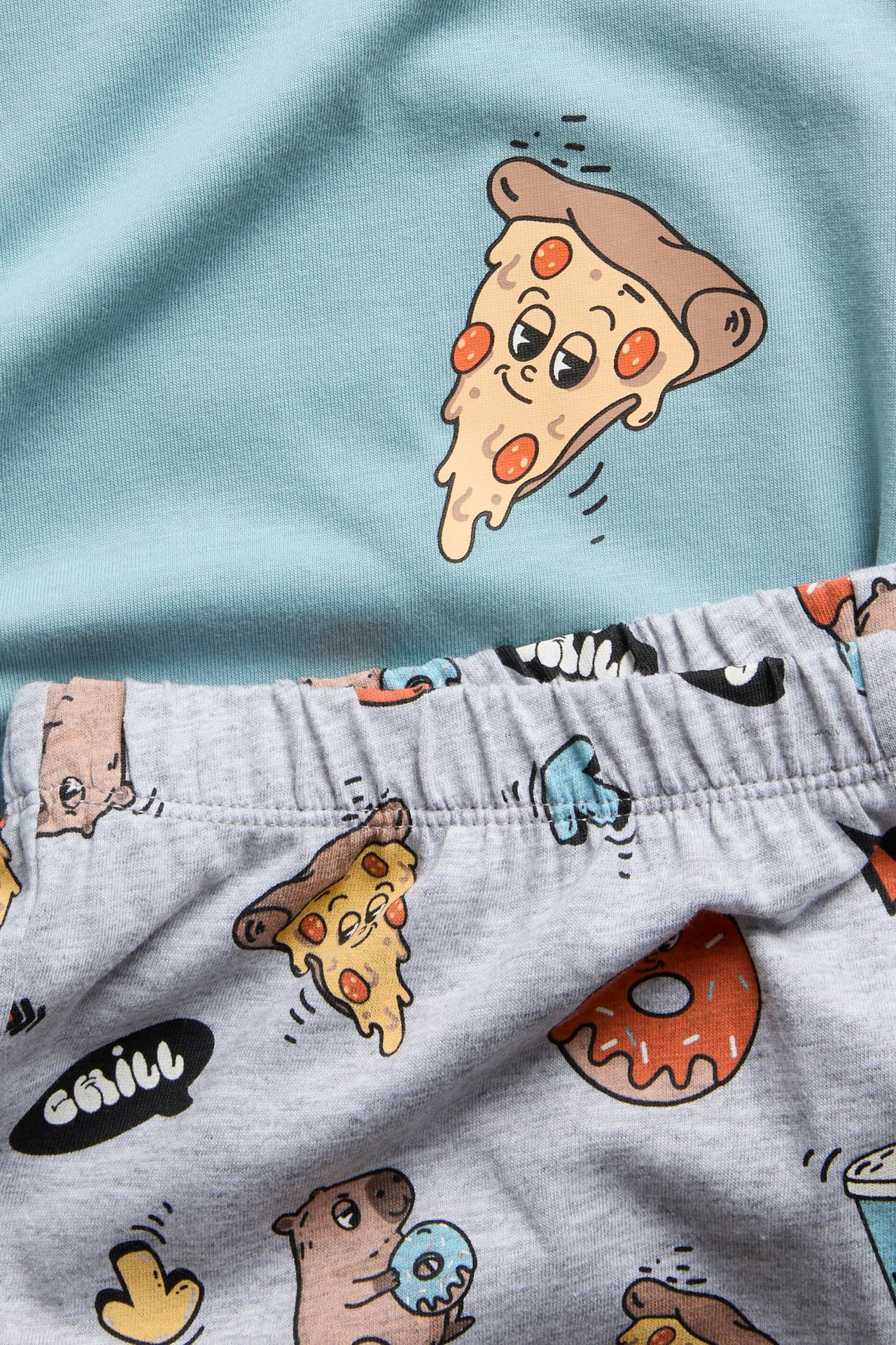 Lähikuva vaaleansinisestä pizzakuvioisesta yläosasta ja harmaista sarjakuvakuvioisista pyjamahousuista.