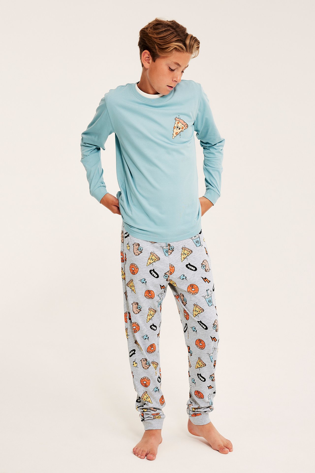Lapsi edestä: vaaleansininen pitkähihainen pyjamapaita pizzakuvalla ja harmaat kuviolliset pyjamahousut.