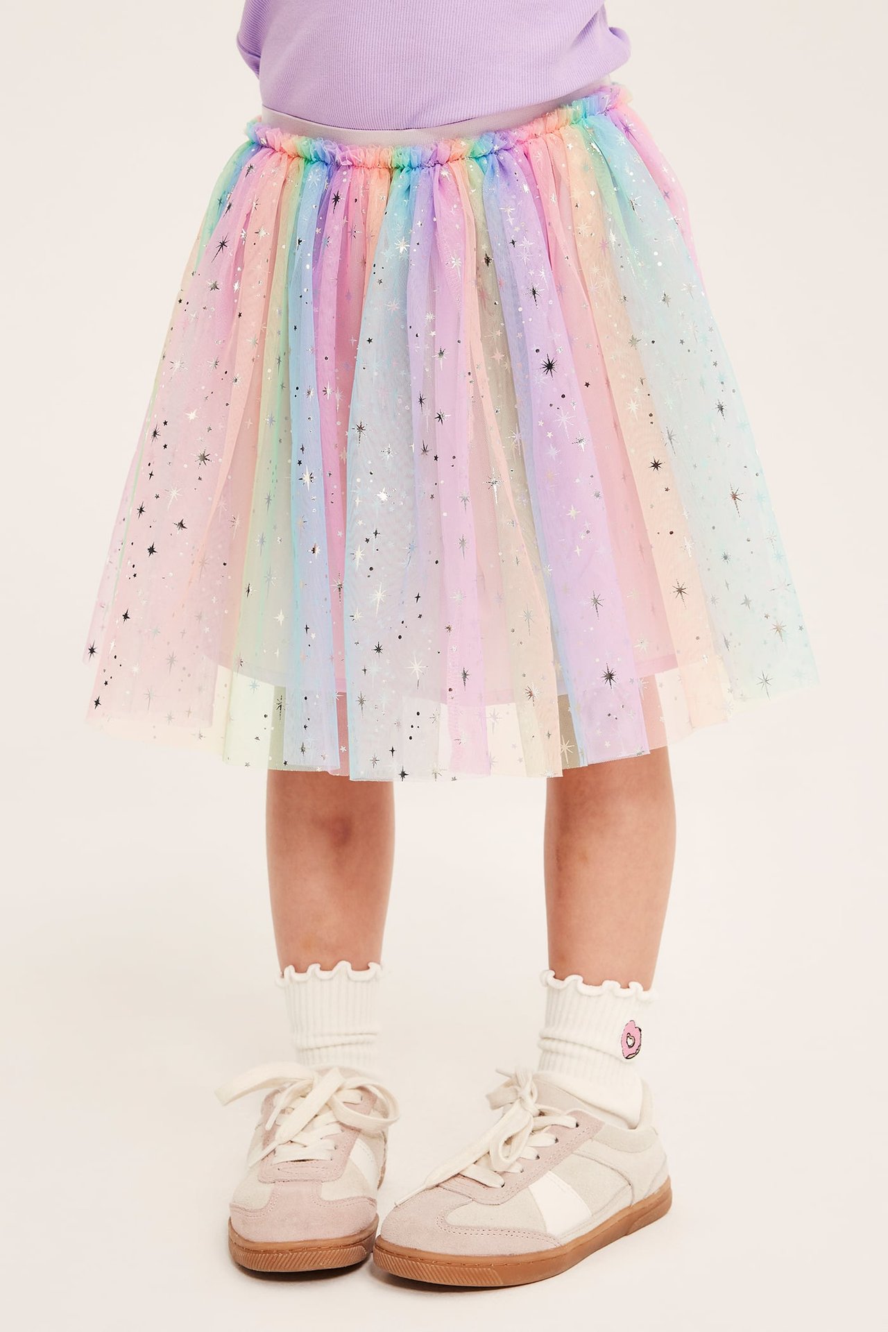 Framifrån: Barn i regnbågsstjärnig tyllkjol, lila topp, volangstrumpor och rosa sneakers.