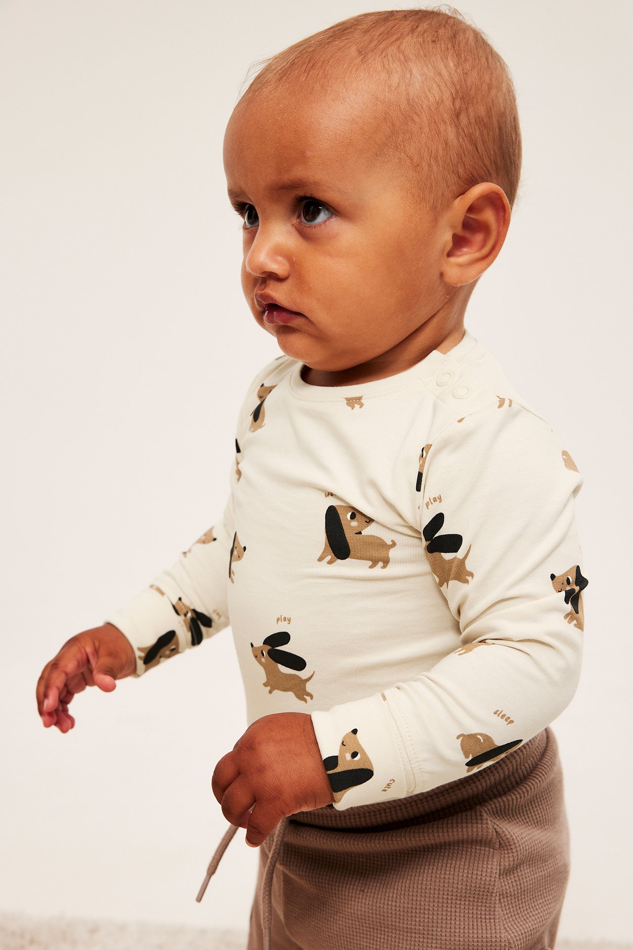 Sivukuva vauvasta off-white pitkähihaisessa koiraprinttipaidassa ja ruskeissa vohvelineulehousuissa.