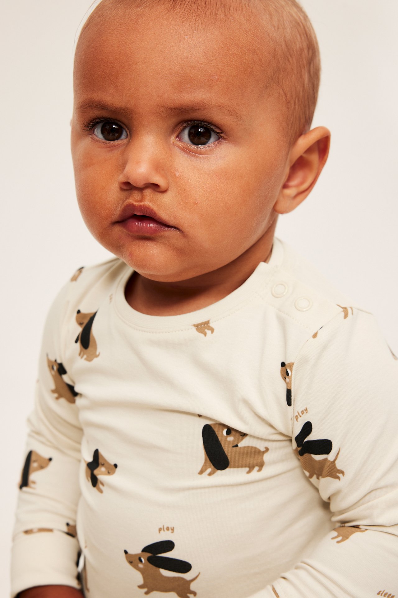 Baby bär en krämfärgad långärmad topp med brunt och svart hundtryck och axelknappar.