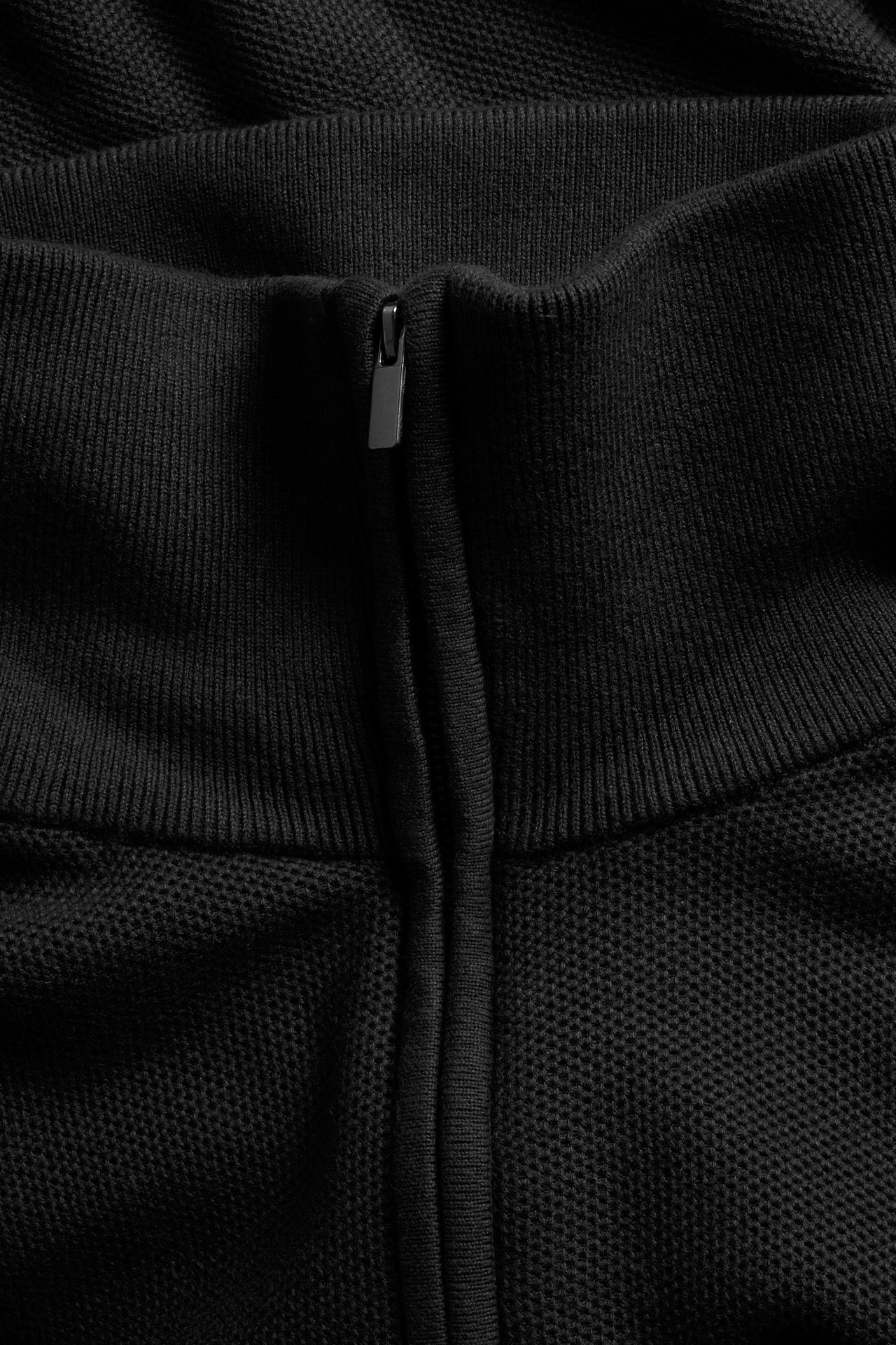 Nærbilde av en svart strukturstrikket topp med ribbestrikkede detaljer og kort glidelås.