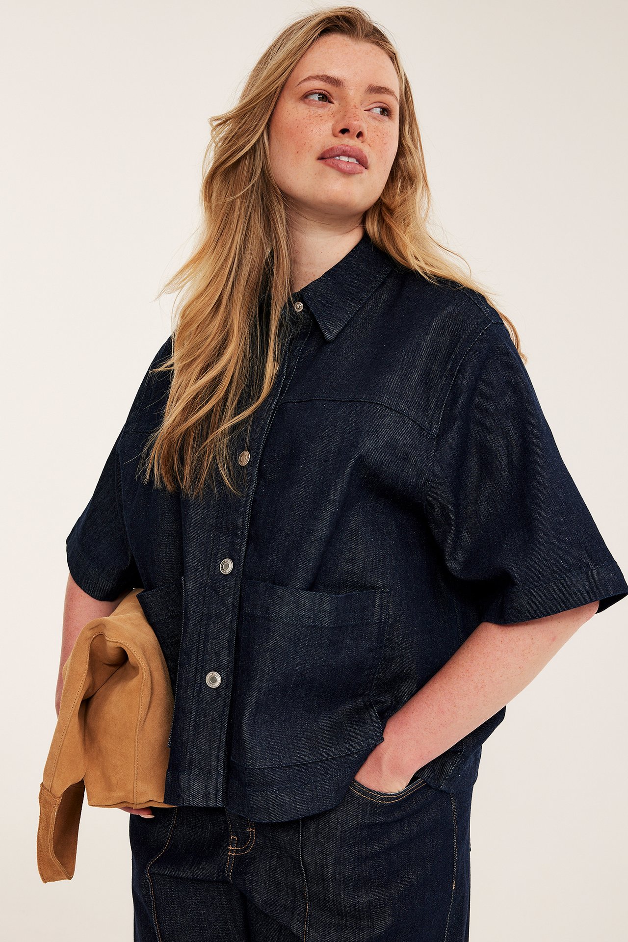 Frontbilde: kvinne i mørkblå denim kortermet skjorte, matchende jeans, holder brun clutch.