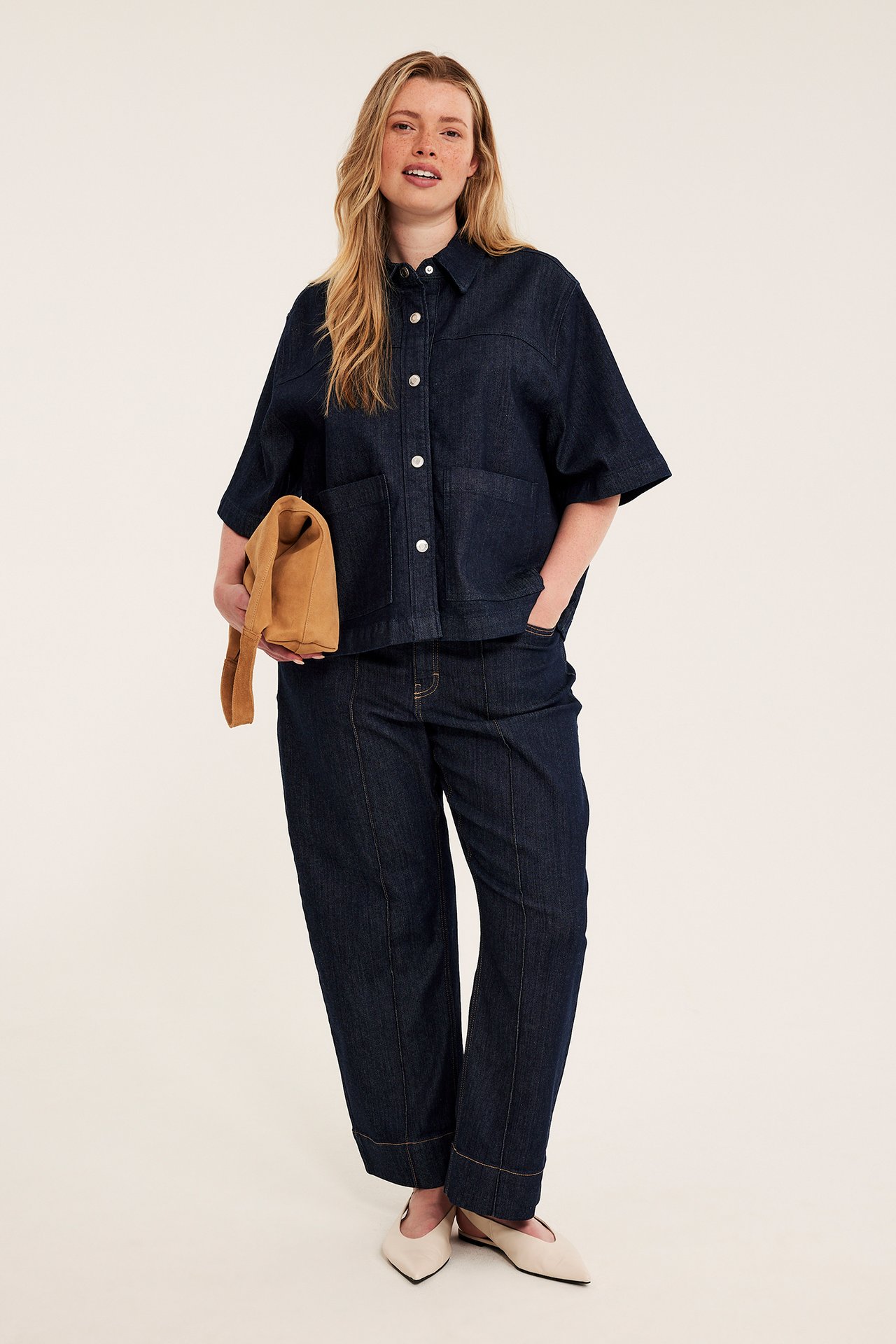 Front: Kvinne i mørkeblå denimskjorte, matchende bukser, holder brun veske, beigefargede sko.