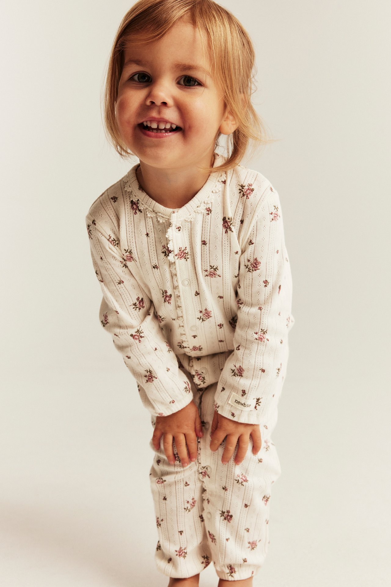 Framifrån: Barn i en krämfärgad blommig och randig hel pyjamas med knappar och krås.