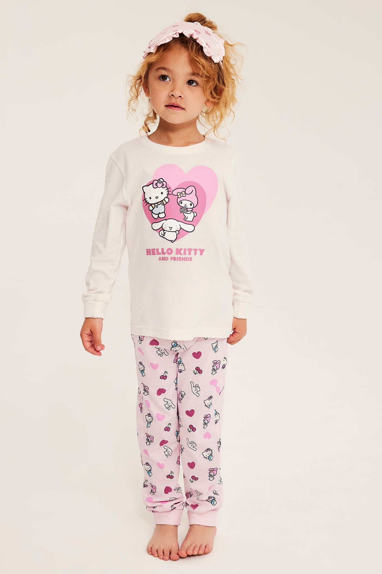 Forfra, et barn i hvit Hello Kitty-pyjamas med trykk og et rosa volangpannebånd.