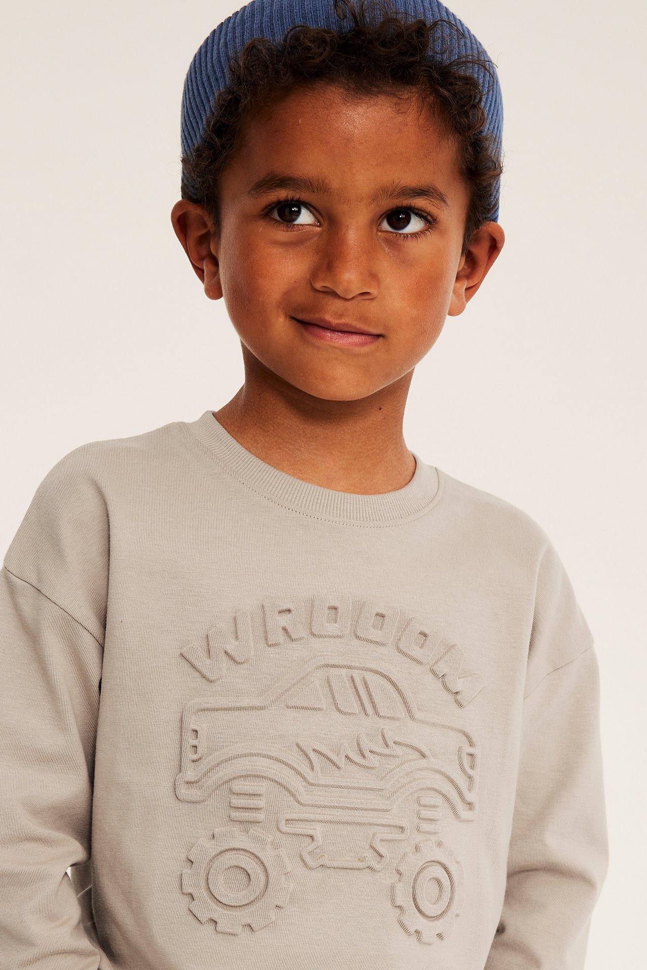 Närbild på barn i ljusbeige sweatshirt med präglad monstertruck och blå mössa.
