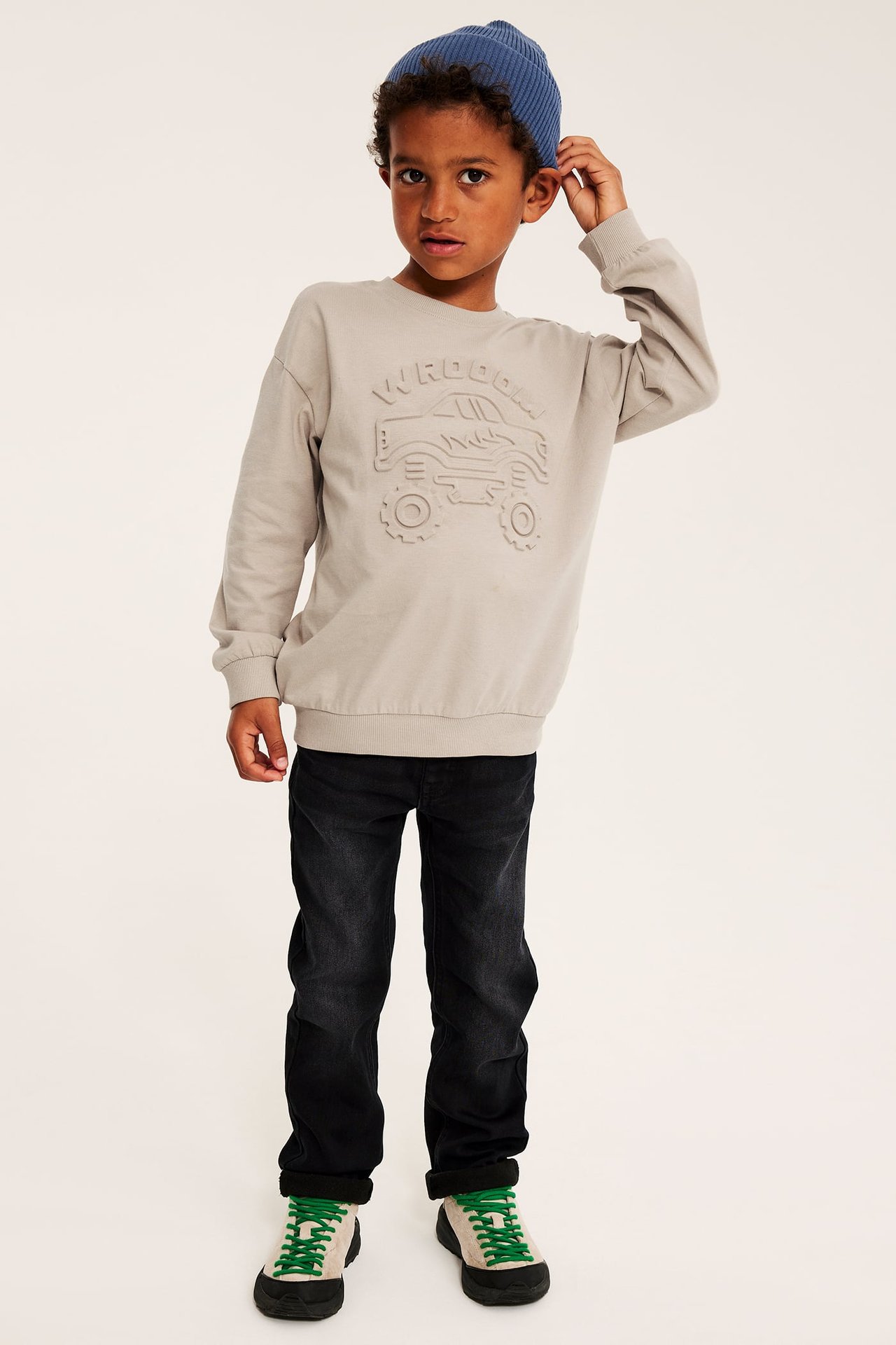 Front view av ett barn i ljusgrå monstertrucktröja, mörka jeans, blå mössa och sneakers.