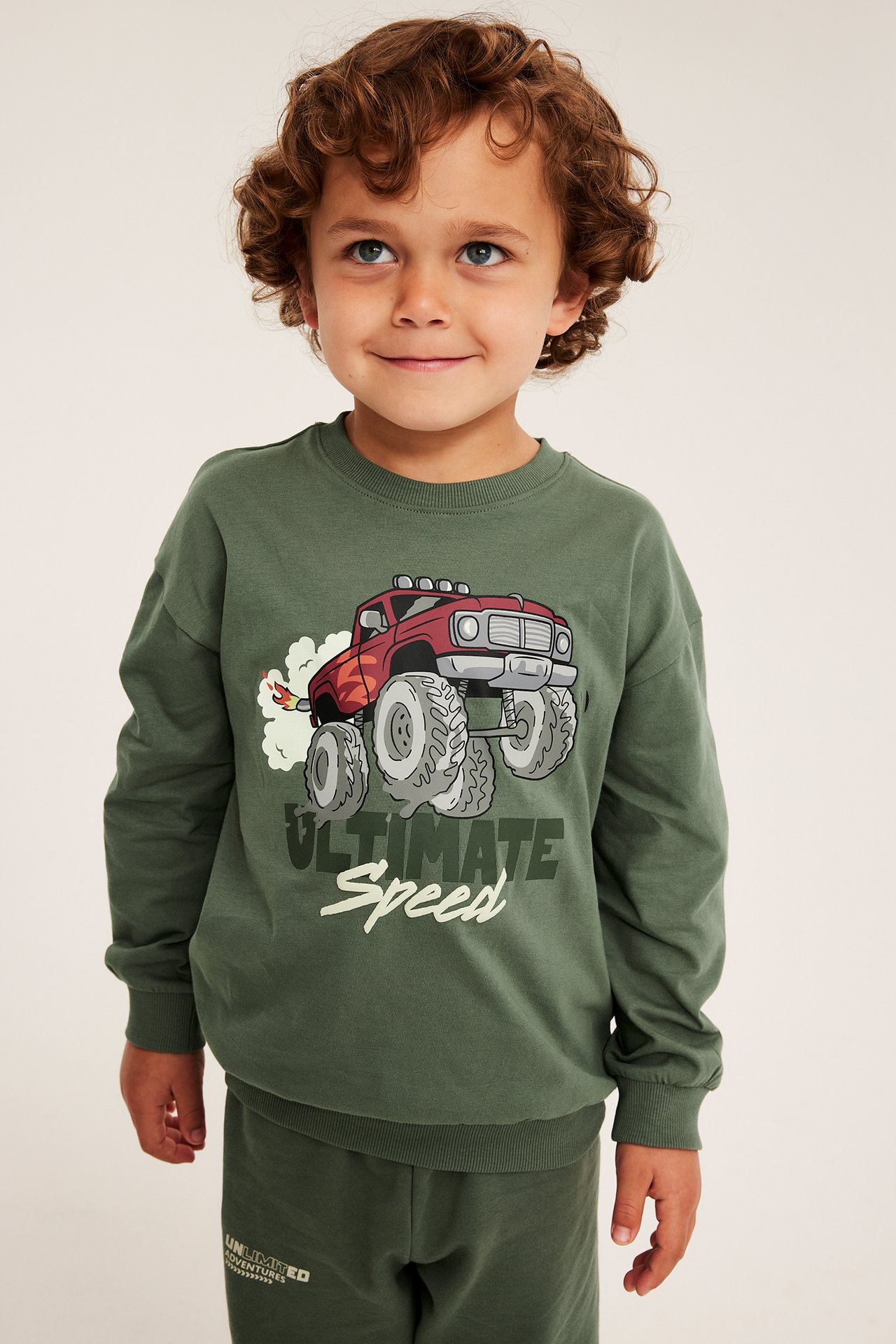 Framifrån, barn i grön långärmad sweatshirt med monstertryck och gröna sweatpants.