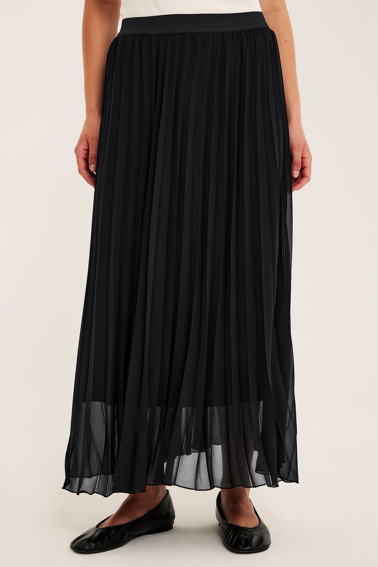 Framifrån: en kvinna bär en lång svart plisserad kjol, en ljus topp och svarta ballerinaskor.