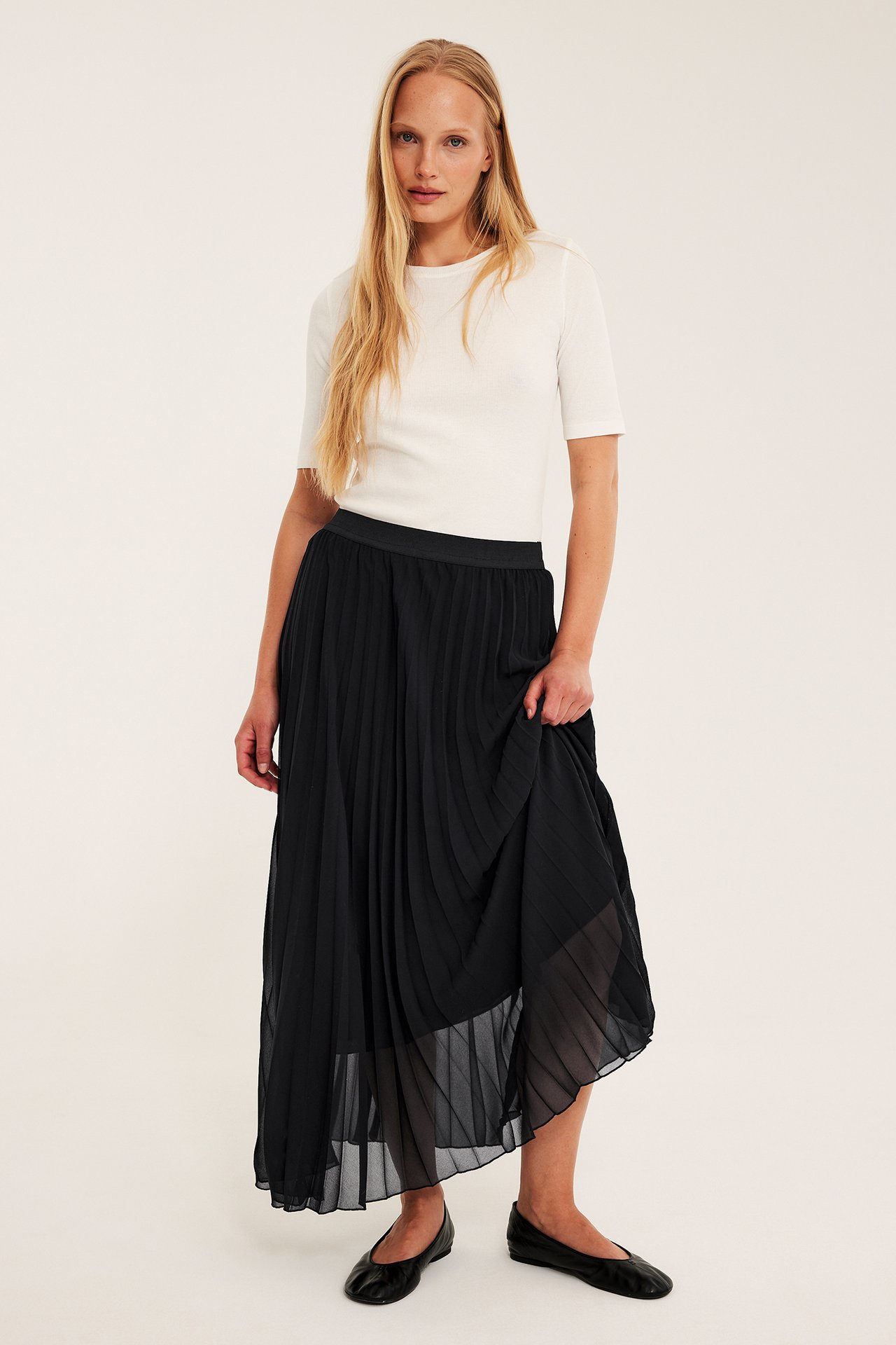 Framifrån: Kvinna i en svart plisserad långkjol, vit kortärmad topp och svarta ballerinaskor.