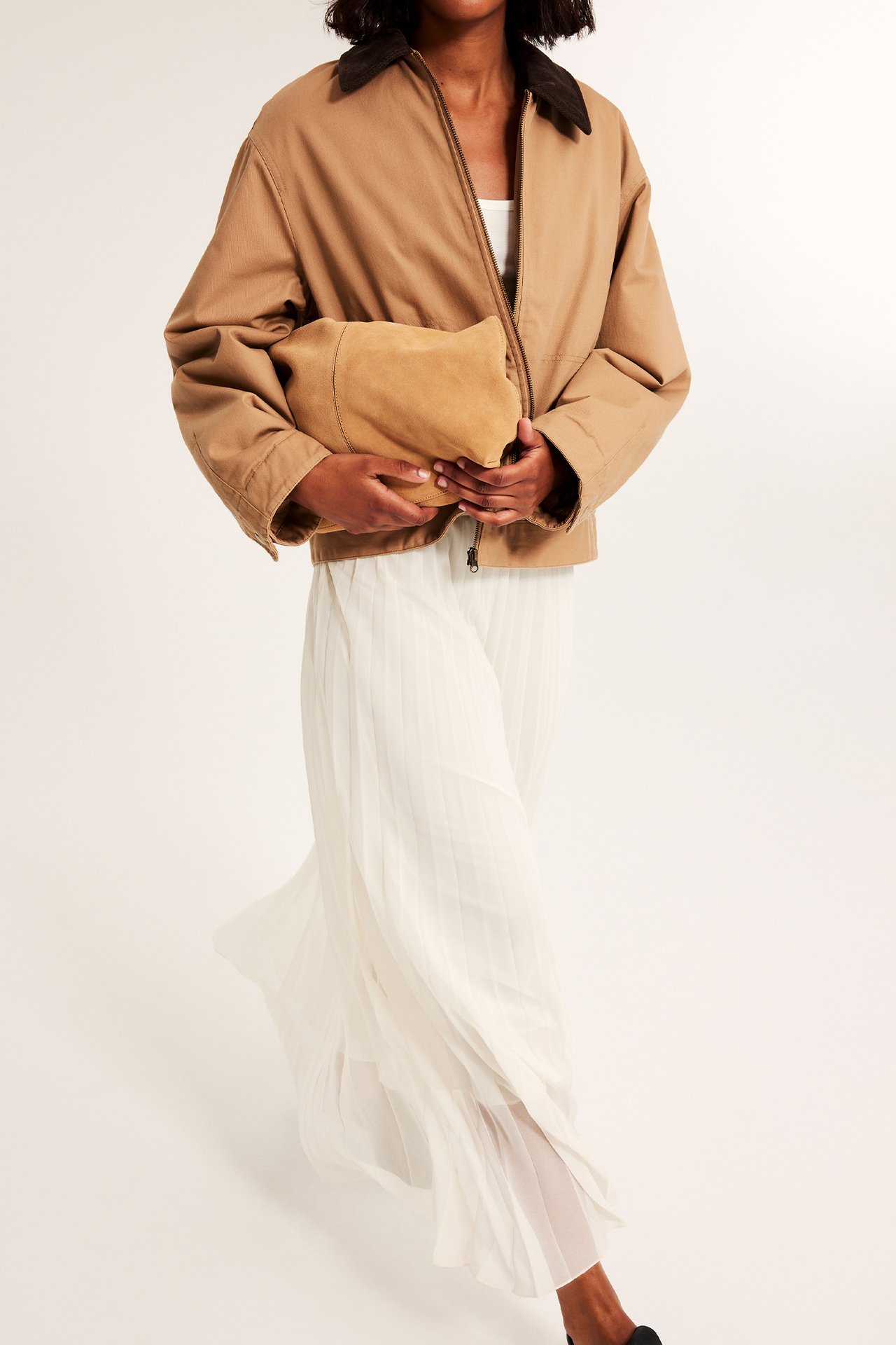 Front vy av kvinna som håller en beige mockaclutch, bär en beige jacka och vit veckad kjol.