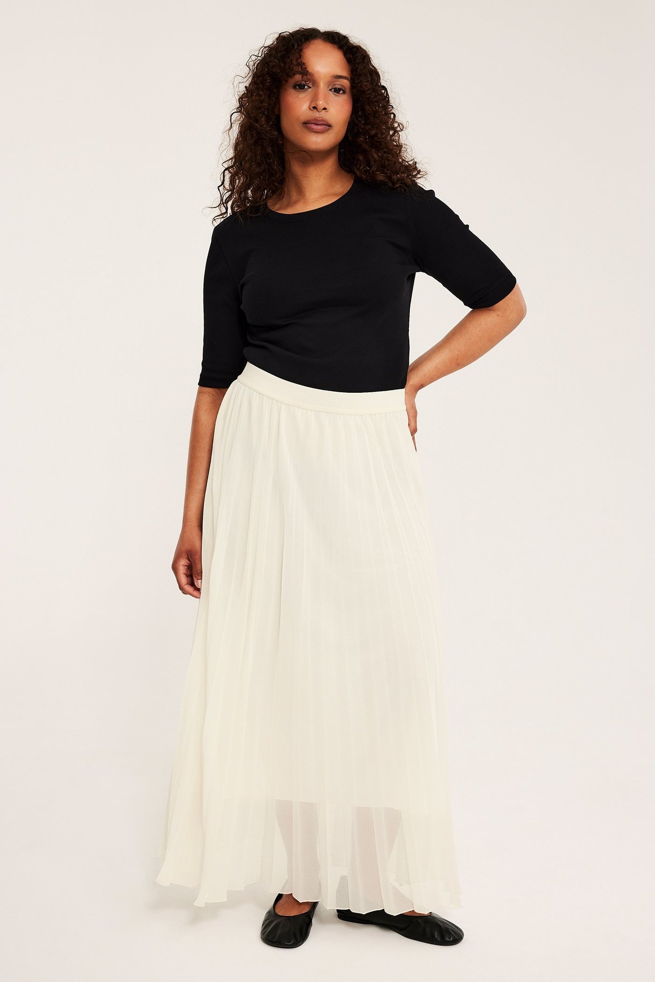 Frontbilde av kvinne i offwhite langt plissert skjørt, svart kortermet topp og svarte ballerinasko.