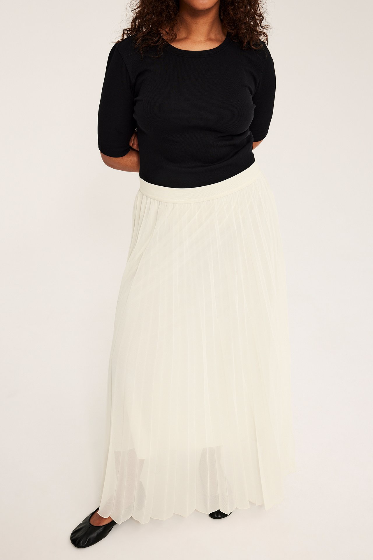 Widok z przodu kobiety w długiej plisowanej spódnicy w kolorze złamanej bieli, czarnym topie i czarnych butach.