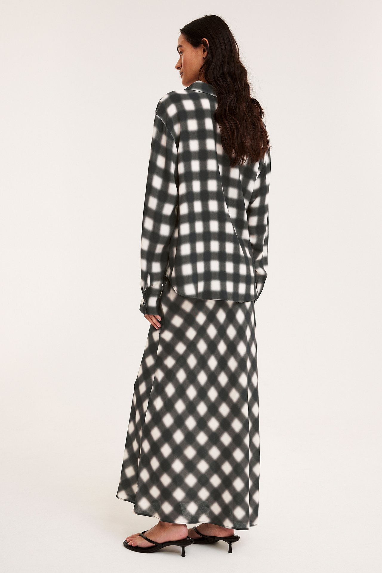Från sidan bakifrån, kvinna i svartvitrutig långkjol av viskos, matchande skjorta och svarta klackar.