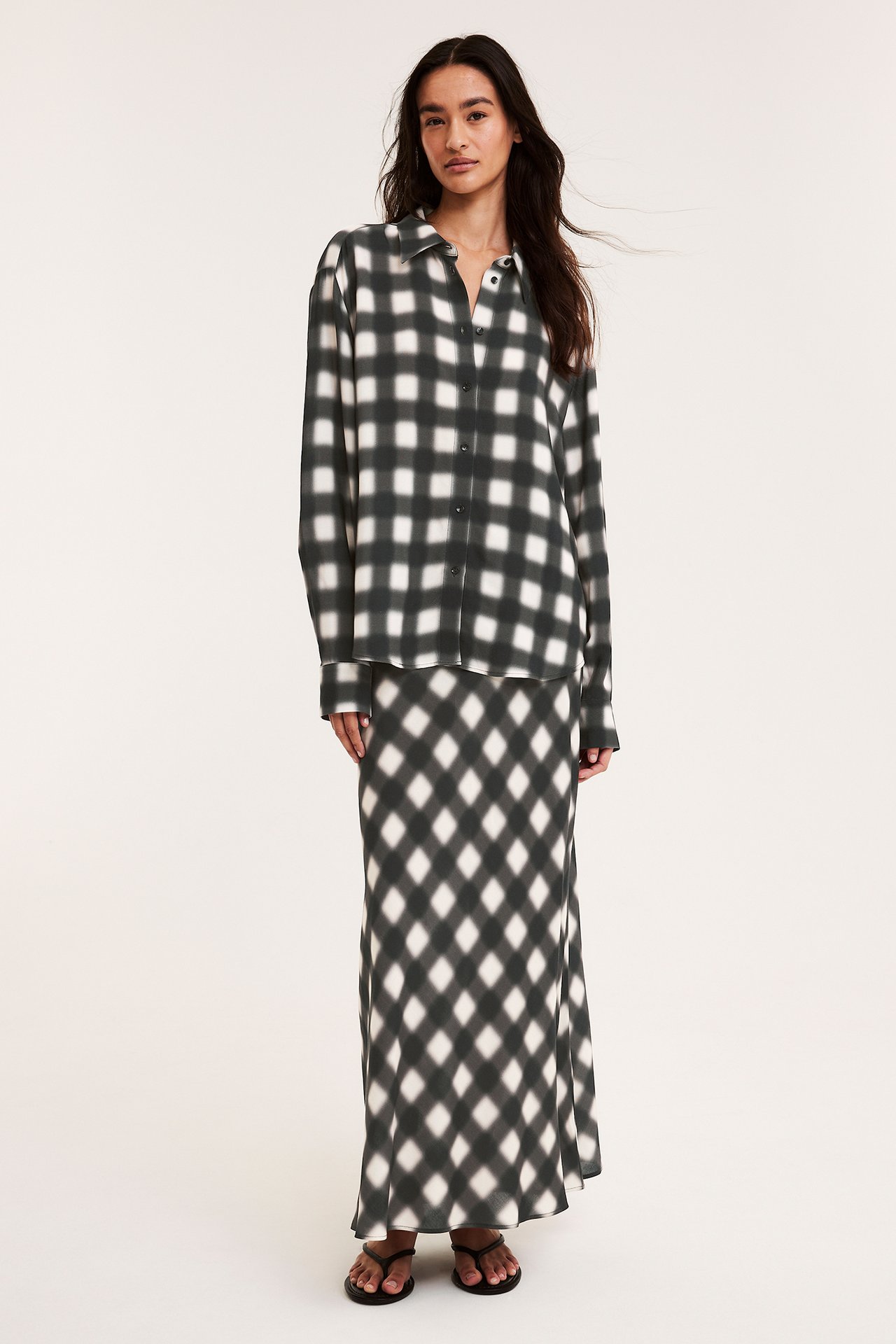 Framifrån: kvinna i svartvit rutig långkjol, matchande långärmad skjorta och svarta flip-flops.