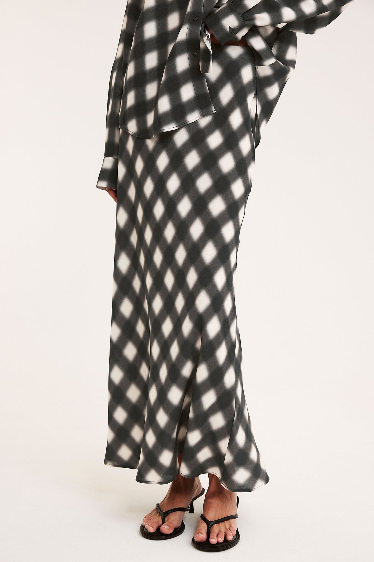 Sidovy av en kvinna i en svartvit rutig viskoslångkjol, matchande skjorta och svarta klackade tåsandaler.