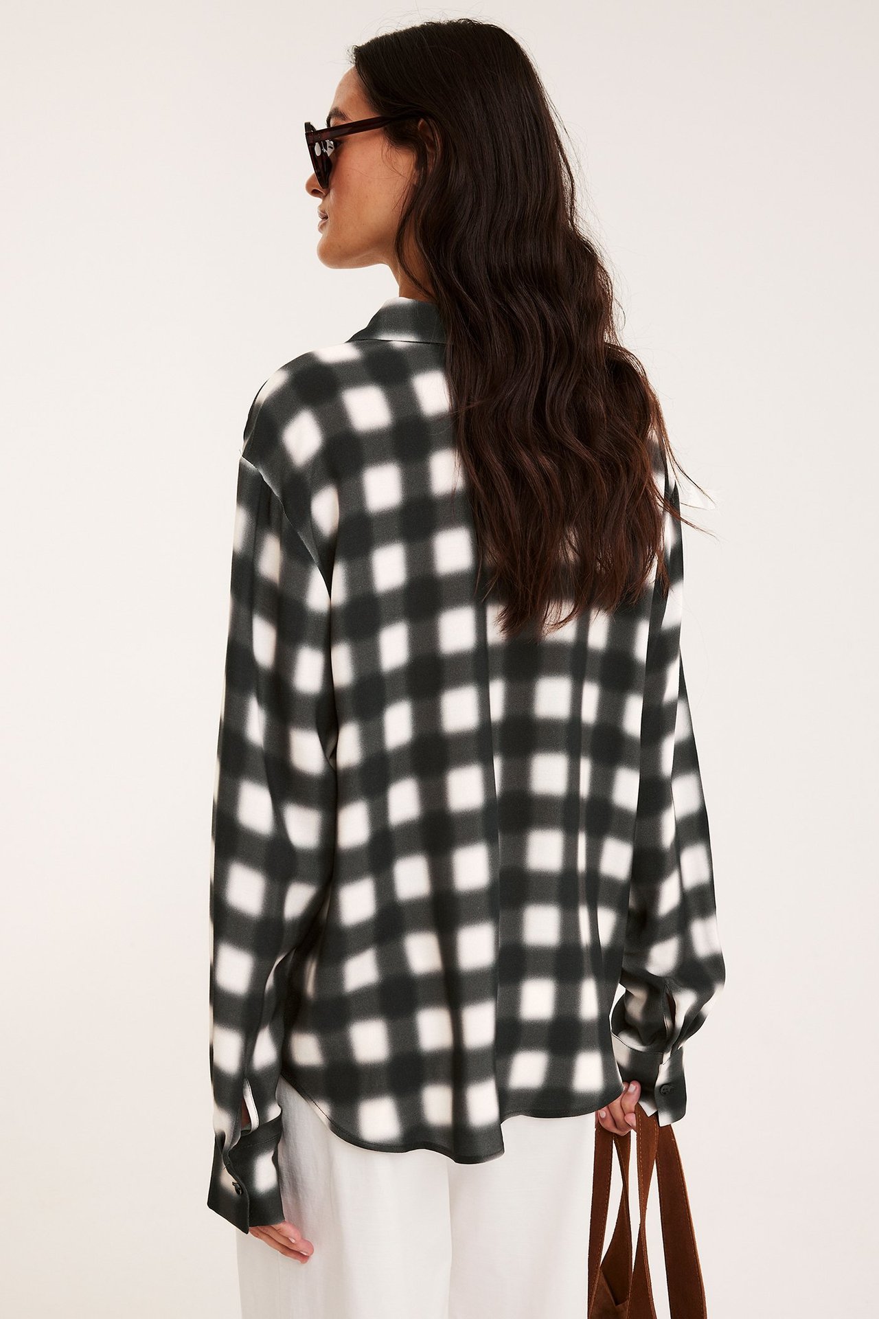 Bakifrån, kvinna i svart- och vitrutig viskosskjorta, vita byxor och mörka solglasögon, håller i brun väska.