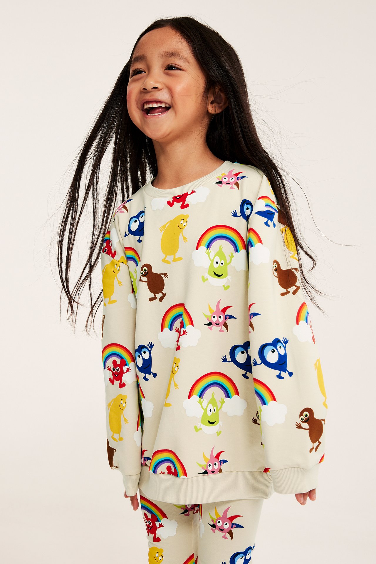 Framsida av ett barn i en beige regnbågs- och figurmönstrad sweatshirt och matchande byxor.