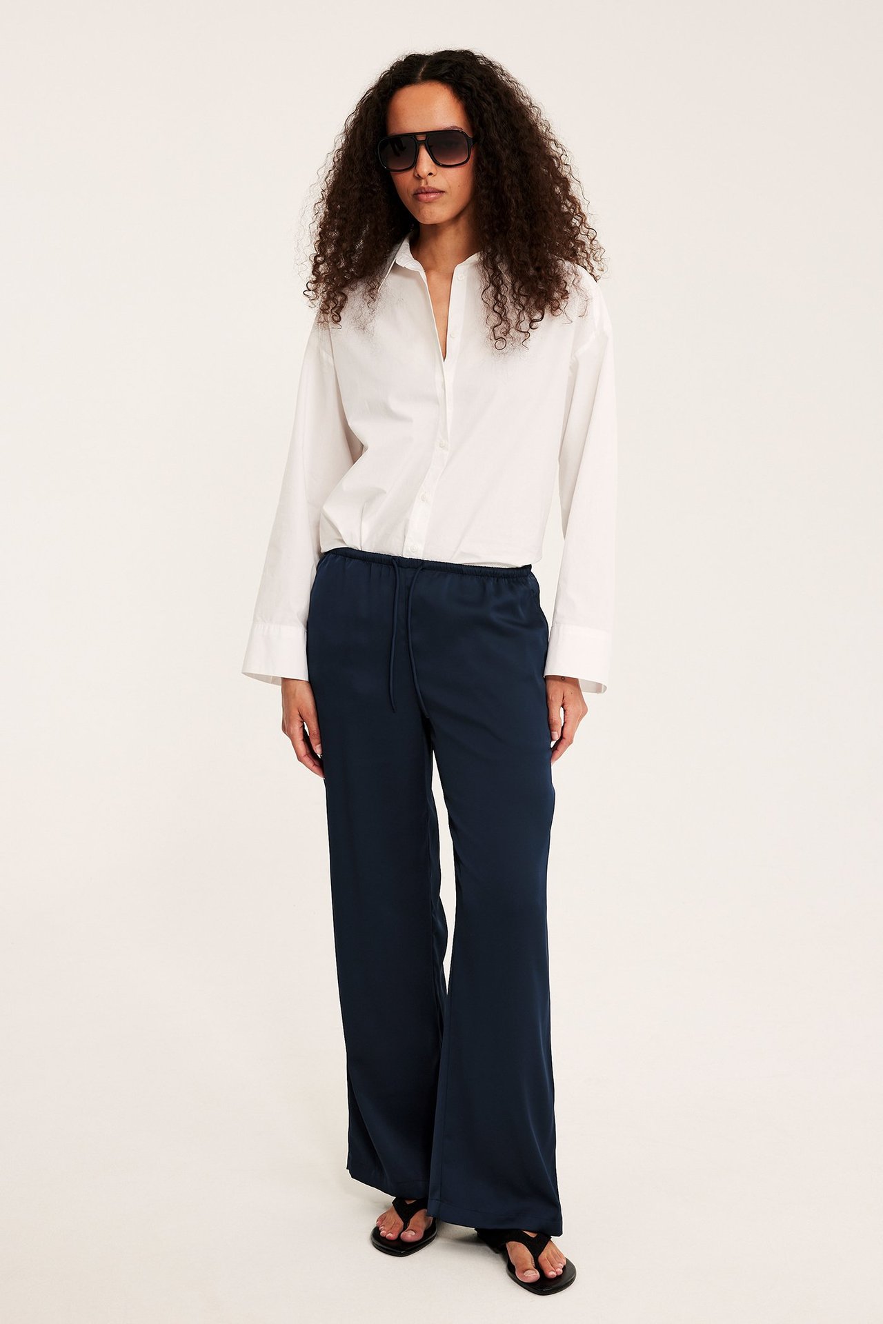 Edestäpäin nainen tummansinisissä satiinihousuissa kiristysnyörillä, valkoisessa paidassa ja aurinkolaseissa.