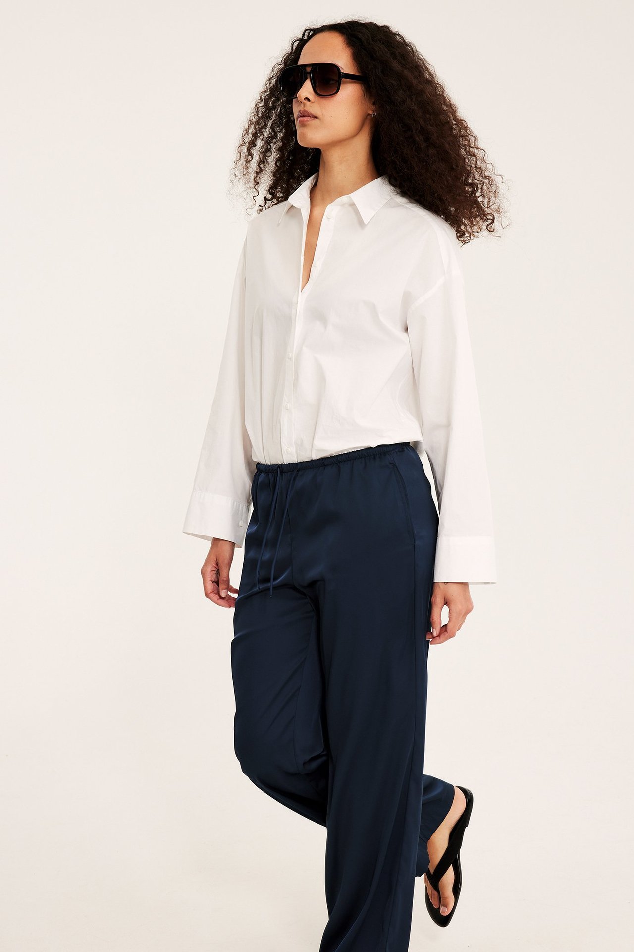Naisen sivukuva tummansinisissä leveälahkeisissa satiinihousuissa, valkoisessa paidassa ja mustissa aurinkolaseissa.