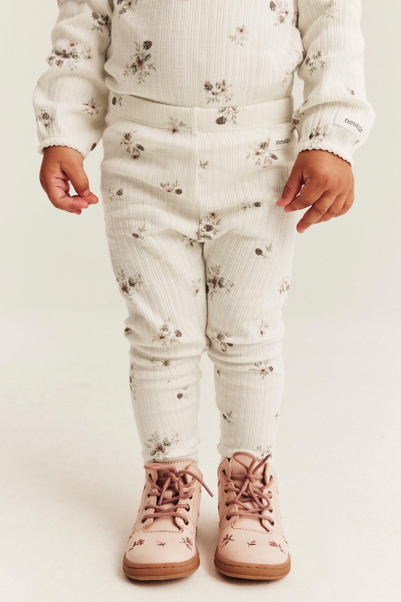 Framifrån, baby i offwhite ribbad pyjamas med blommigt tryck och rosa snörskor.