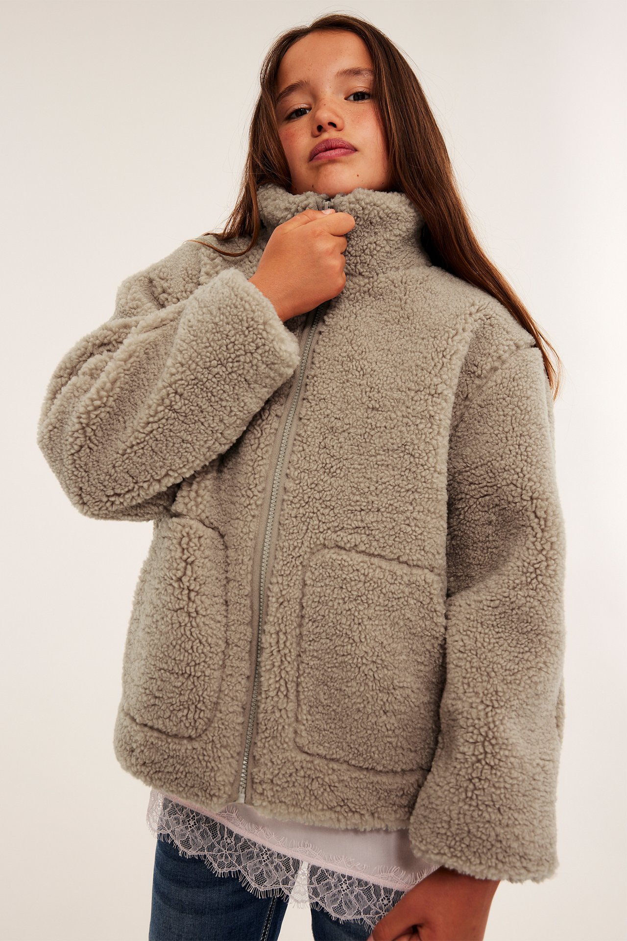 Framifrån, barn i grå fluffig jacka med dragkedja över vit spetstopp och blå jeans.